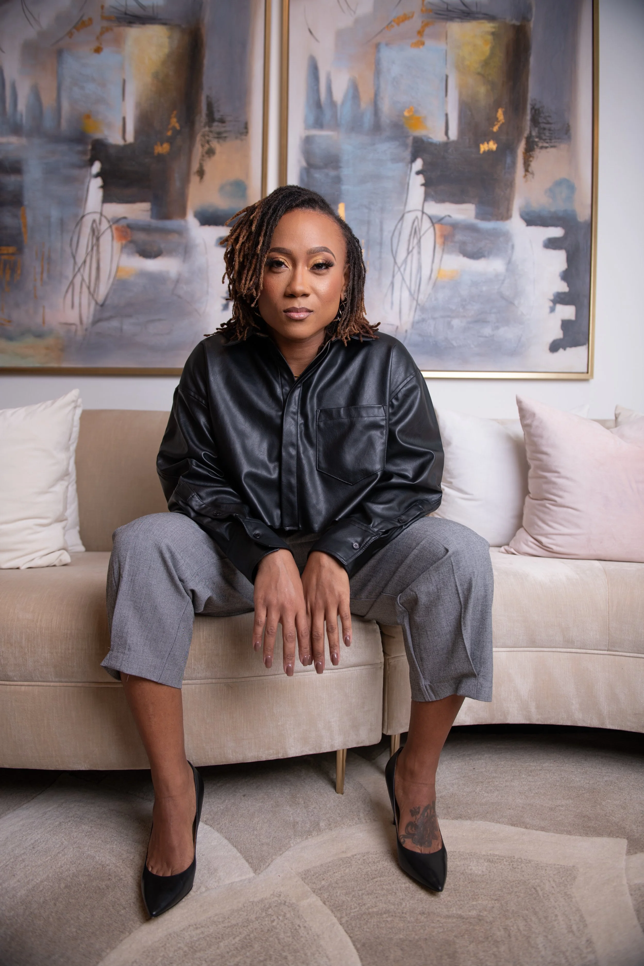 A woman with dreadlocks sitting on a beige couch in front of abstract artwork, wearing a black leather shirt, gray pants, and black high heels.