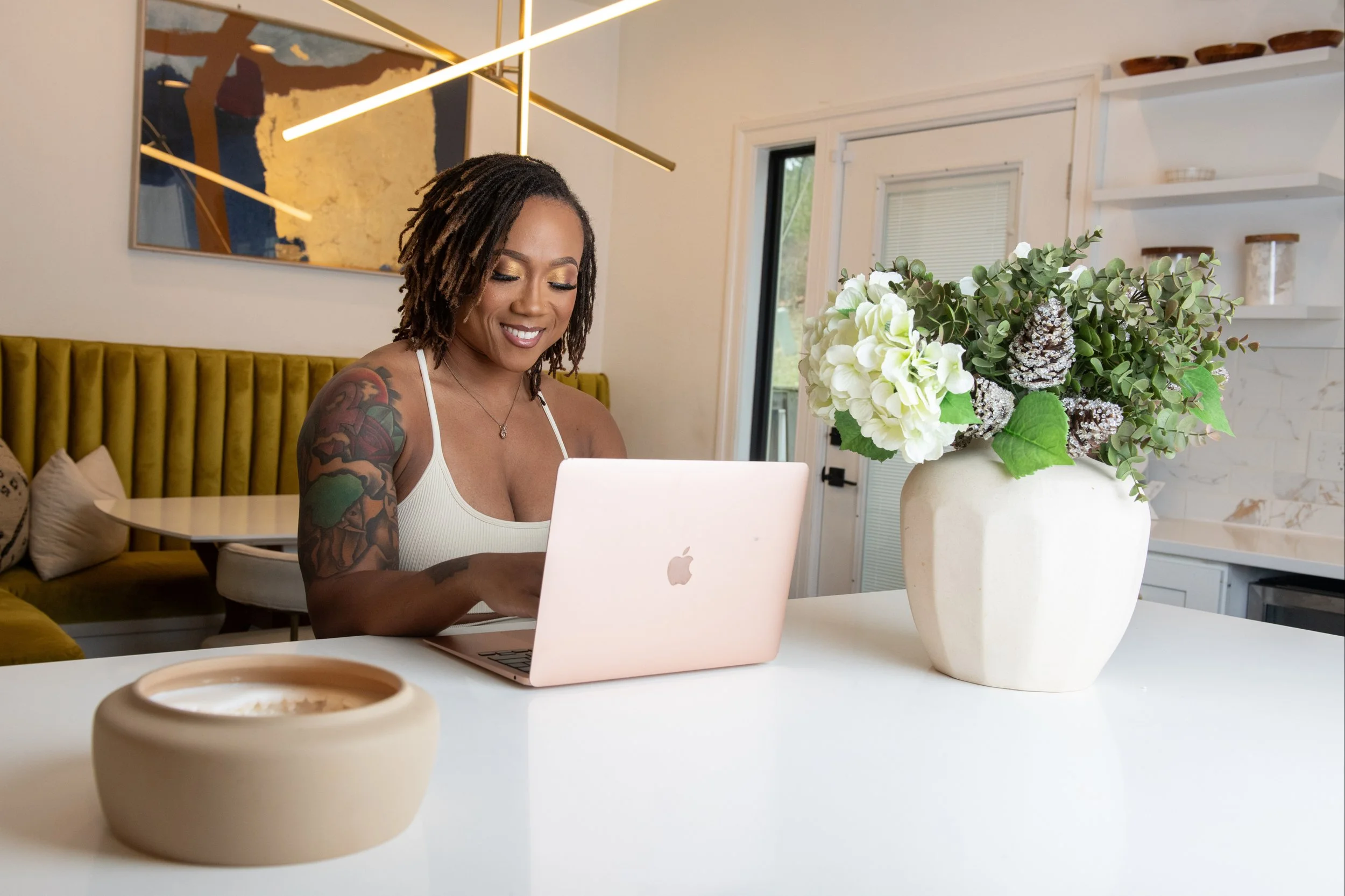 A woman with tattoos on her arm smiling while using a pink MacBook at a white table in a modern kitchen or dining area. There is a large white vase with green and white flowers on the table.