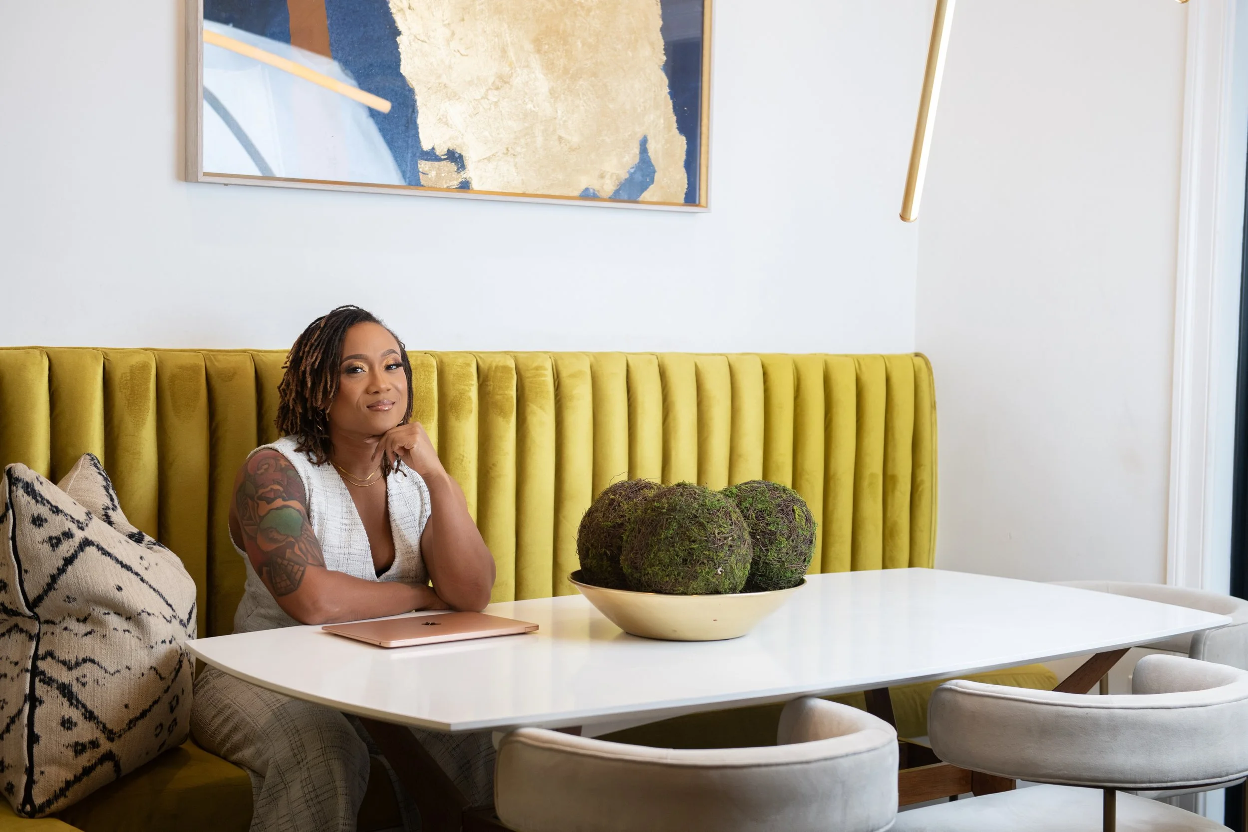 A woman with tattoos on her arm is sitting at a white table with a beige laptop, resting her chin on her hand. She is in a room with white walls, a yellow upholstered bench, a decorative pillow, and a bowl of moss-covered spheres on the table. There is abstract art hanging on the wall behind her.