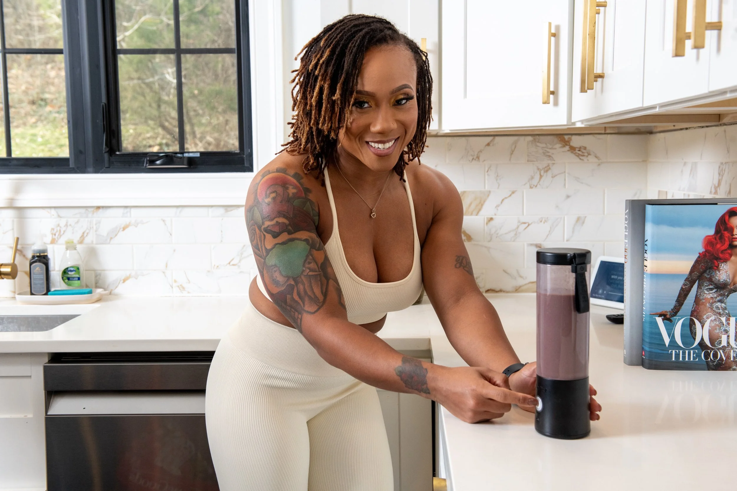 A woman with tattoos on her arms is smiling and operating a blender in a modern kitchen with white cabinets and a marble backsplash.