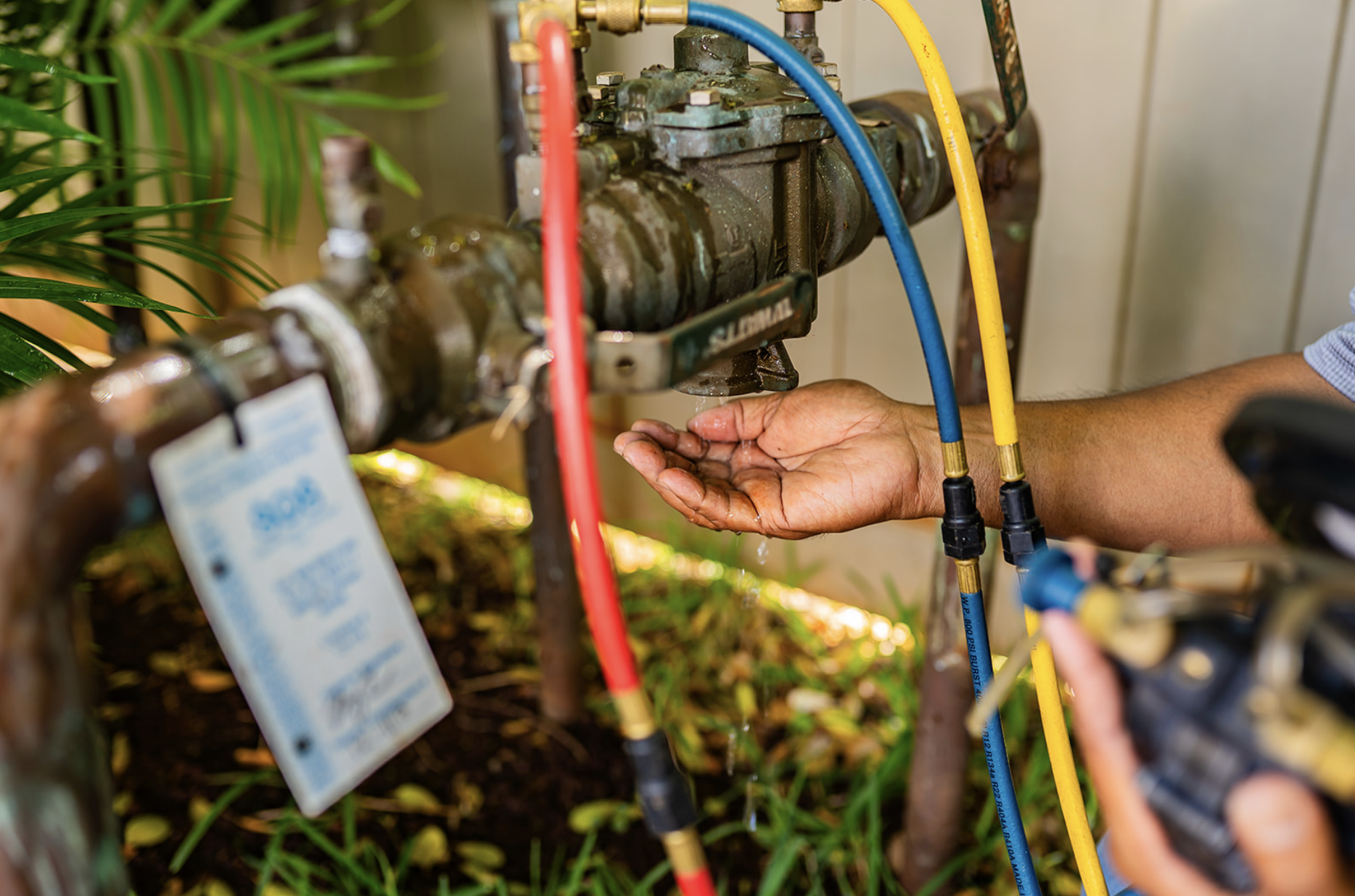 Person testing backflow valve with multiple colored hoses attached, outdoors near greenery.
