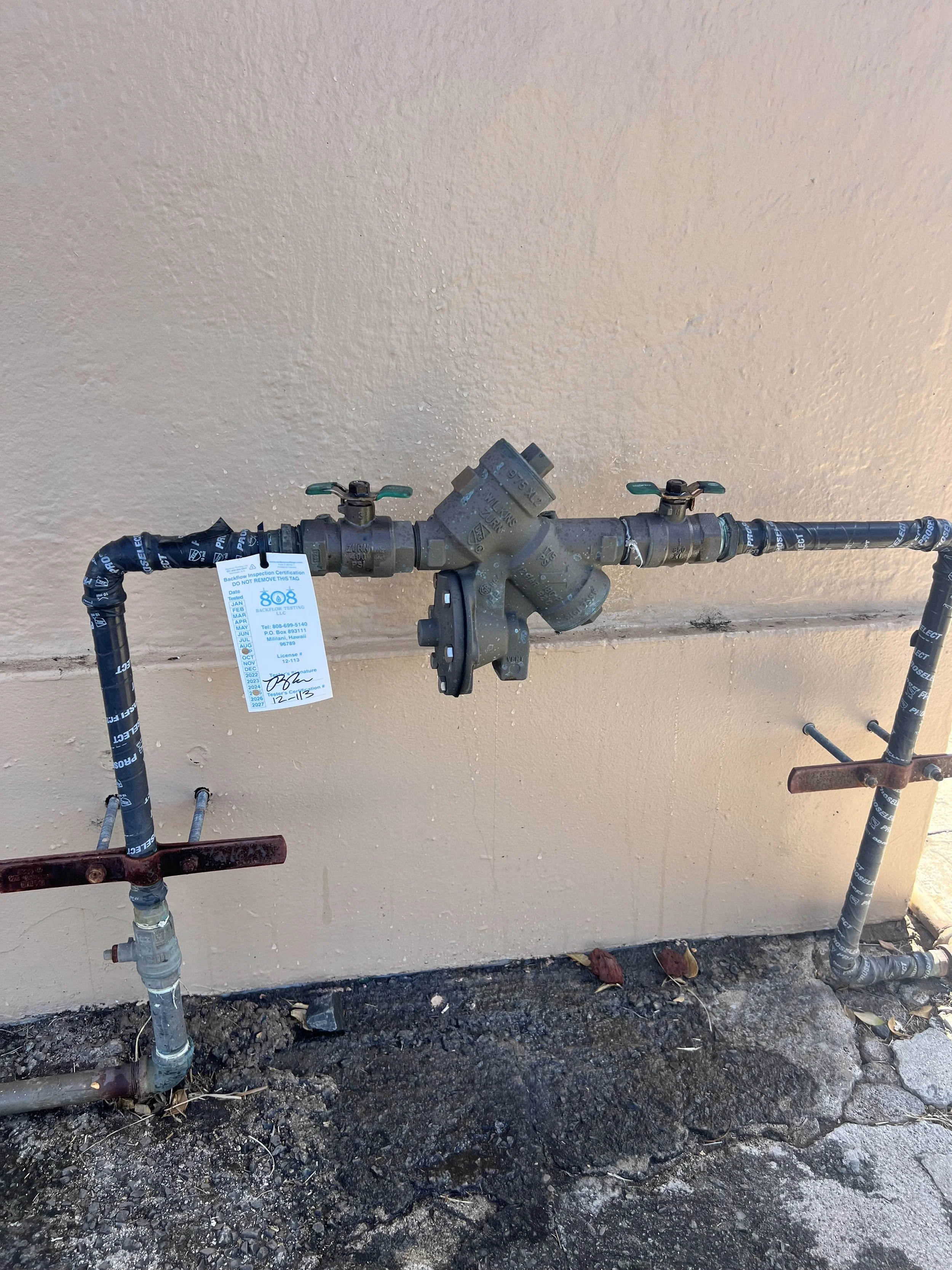 Outdoor backflow device with mounted on a beige wall, with soil and debris on the ground beneath.