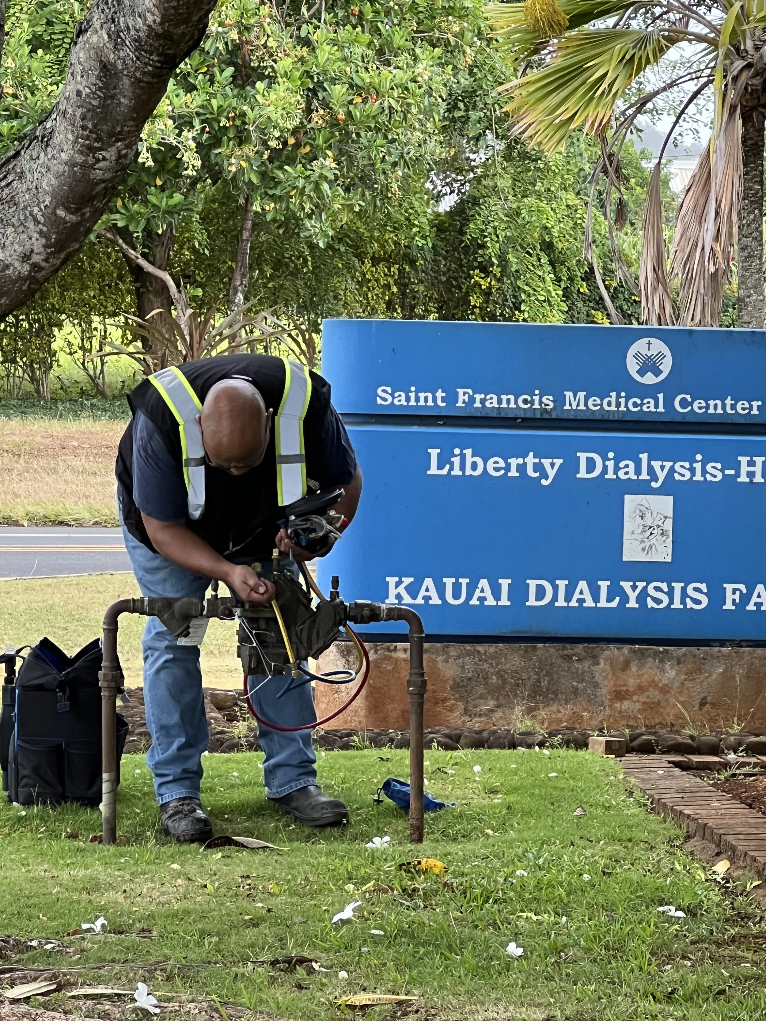 Bert wearing a high-visibility vest and jeans is working on irrigation pipes in front of a blue sign that reads 'Saint Francis Medical Center, Liberty Dialysis, Kauai Dialysis Facility'.