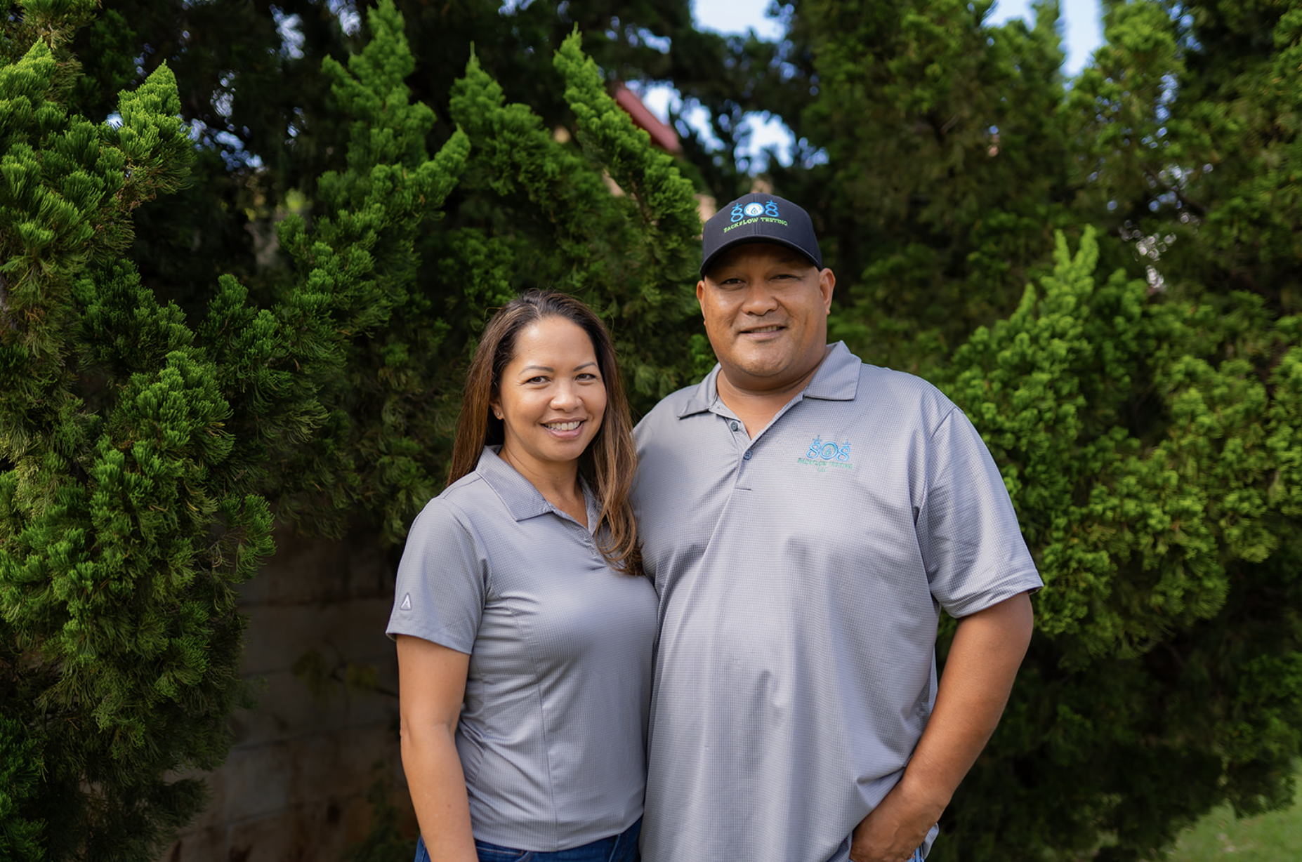Dee and Bert smiling standing outdoors in front of green trees, both wearing gray shirts with 808 backflow testing logo.