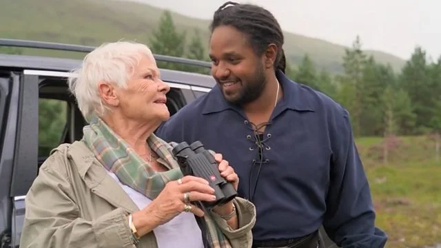 Dame Judi Dench holding binoculars and smiling at Hamza Yassin in a misty Scottish Highland landscape, illustrating the concept of guided vision and nature observation.