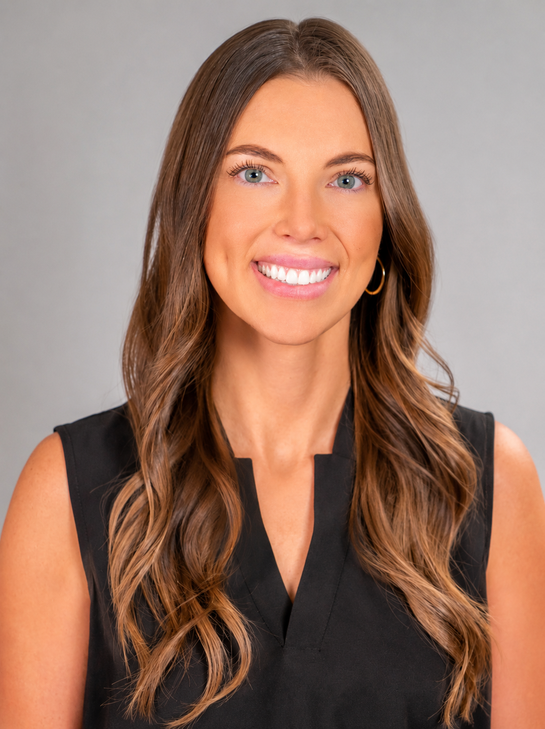 A smiling woman with long wavy brown hair wearing a black sleeveless top and hoop earrings against a light gray background.