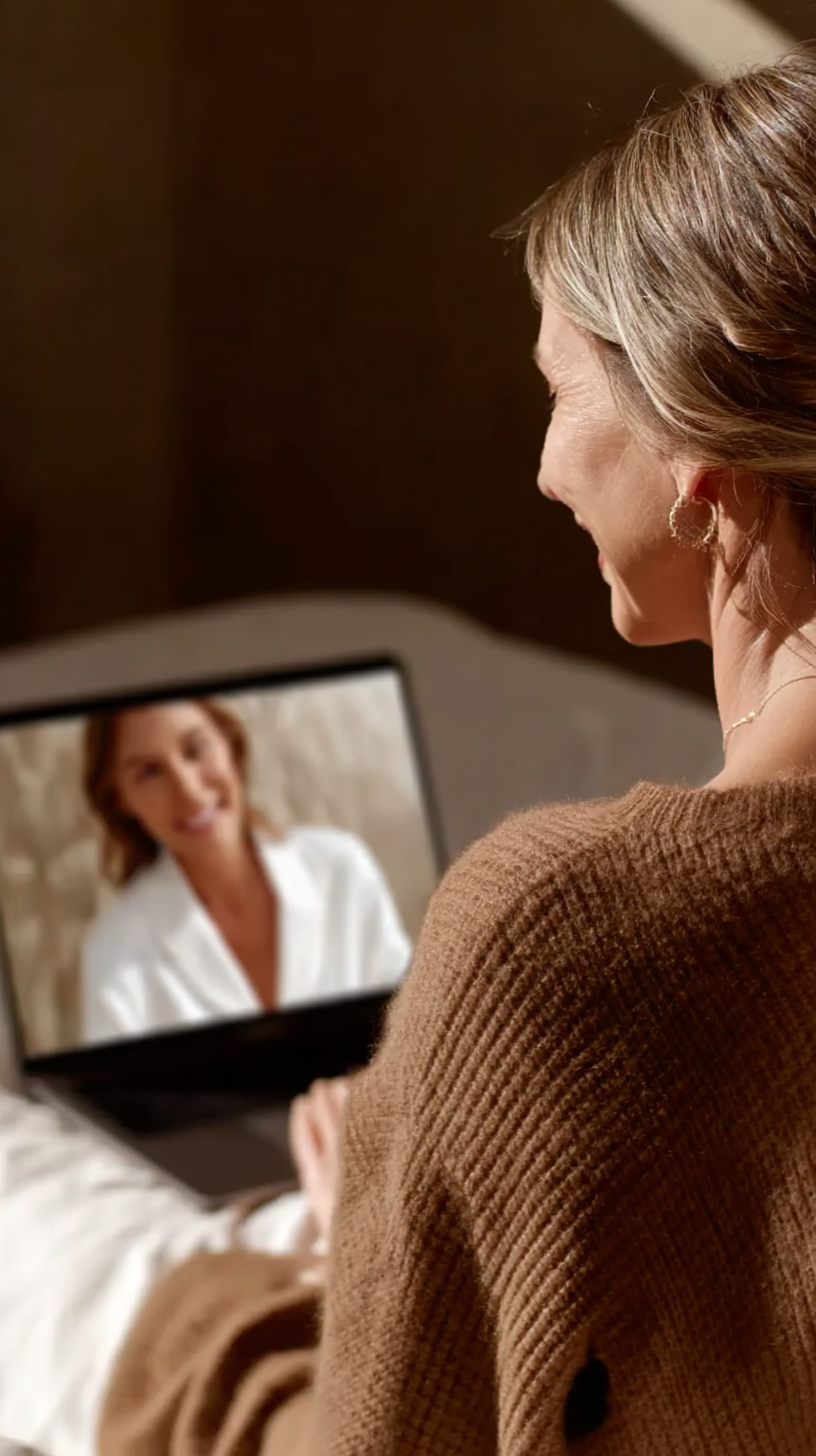 A woman with short blonde hair and earrings is smiling and talking to another woman on a video call on her laptop. The woman on the screen has brown hair, is wearing a white top, and is also smiling.