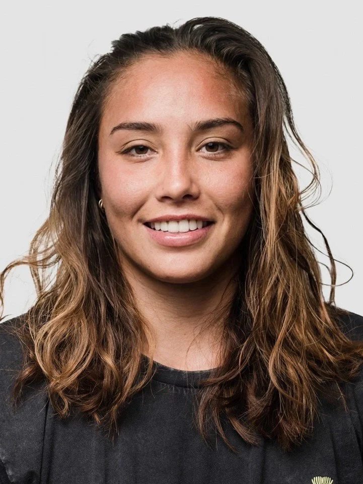 A young woman with wavy brown hair, smiling, wearing a black shirt and small earrings, against a plain light background.