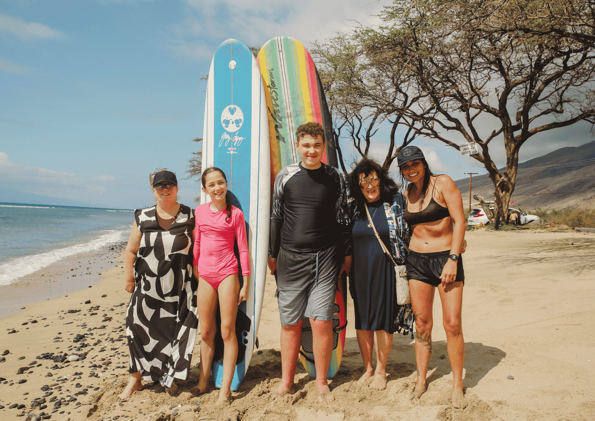 Group of five people standing on a sandy beach, smiling and posing for a photo with surfboards behind them. They are wearing beach and swimwear, with ocean waves and a blue sky in the background.