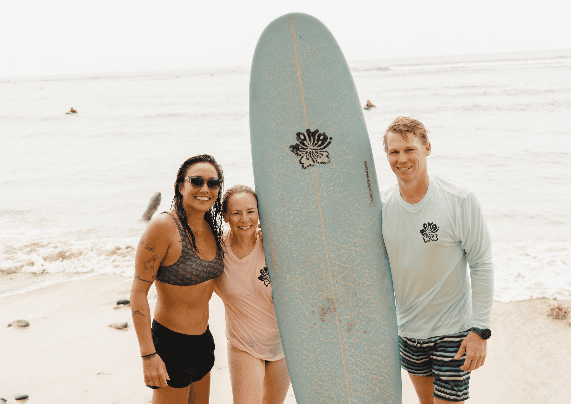 three people at the beach, two women and one man, standing together with an upright surfboard that has a butterfly decal, smiling at the camera, with the ocean and a few surfers in the background.