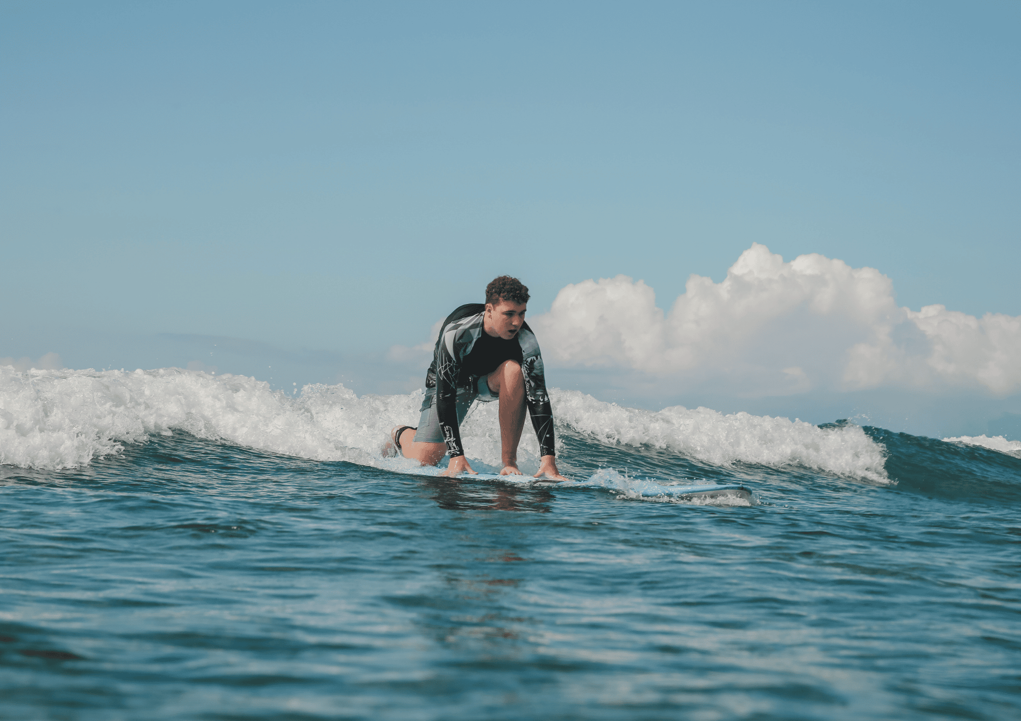 Young man in a black rash guard and gray shorts on a surfboard balancing on a wave under a cloudy sky.