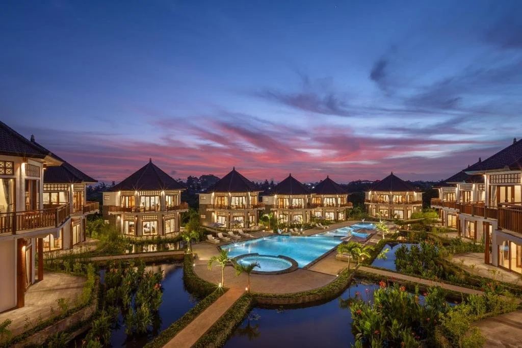 Luxury resort with multiple bungalow-style villas surrounding a large pool, lush landscaping, and a colorful sunset sky.