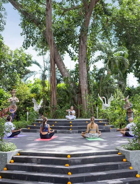 People practicing yoga outdoors on tiered stone steps surrounded by greenery, large trees, and decorative sculptures.
