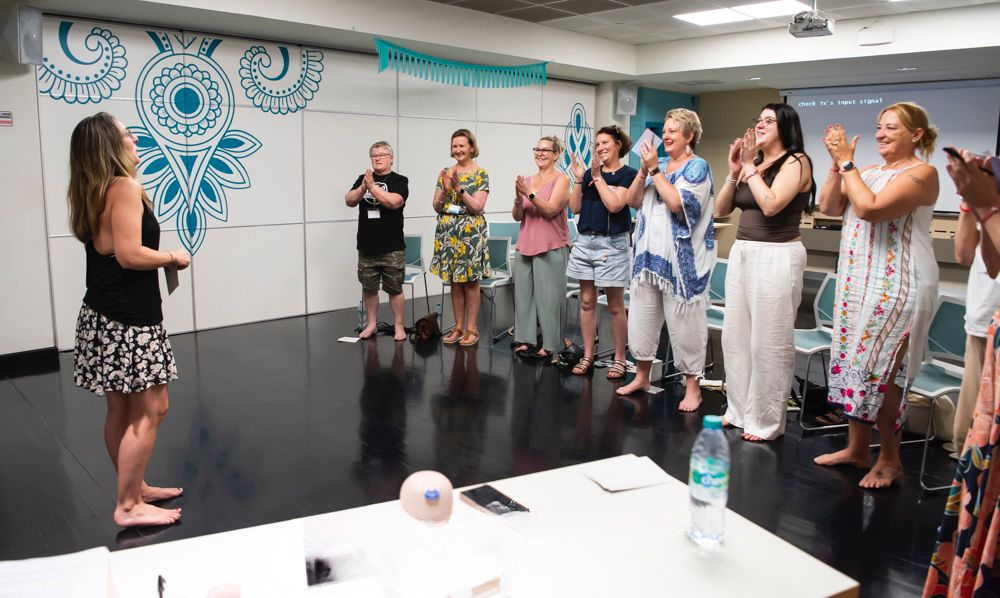 A group of women standing in a room with a dark floor and white walls, many of them are clapping and smiling while one woman faces the group.