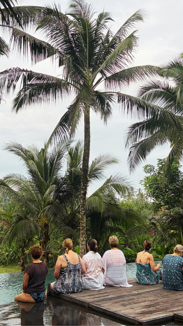 A group of people sitting on a wooden platform near a pool, facing lush tropical palm trees and greenery under a cloudy sky.
