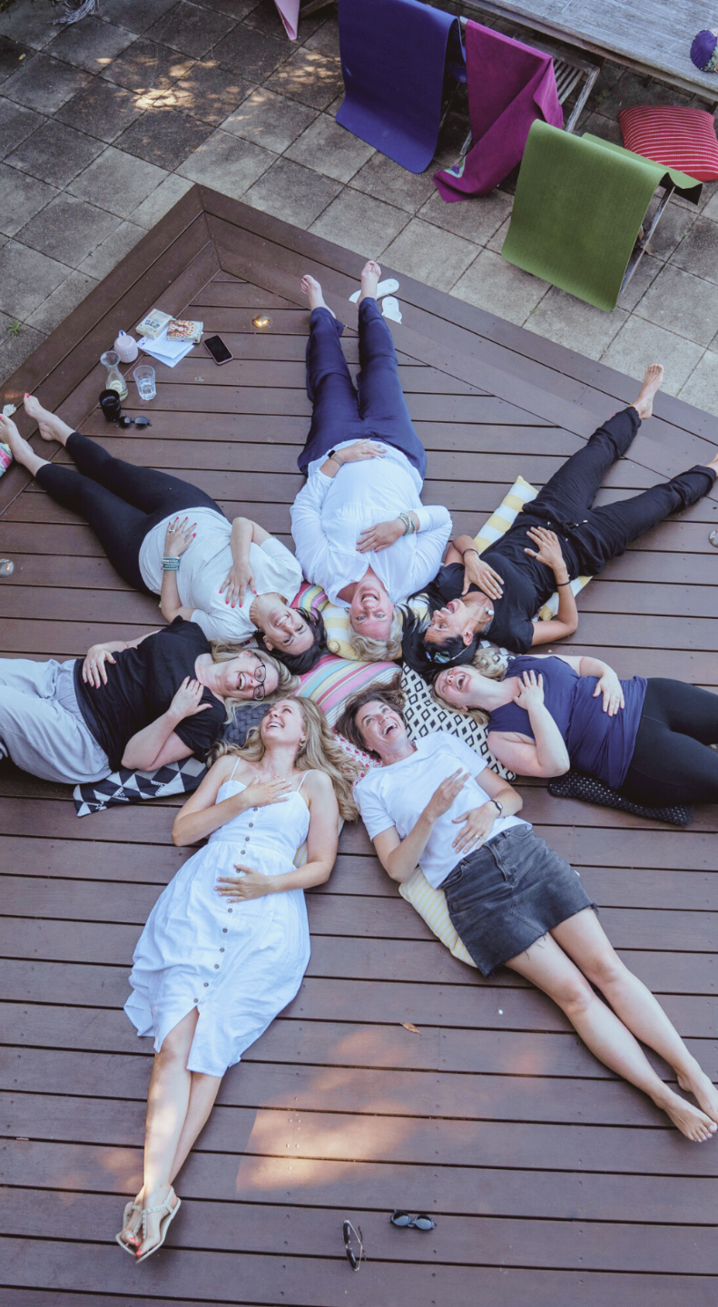 Six women lying on a wooden deck in a circle, smiling and laughing, with some resting on pillows, surrounded by drinks and snacks in an outdoor patio setting.