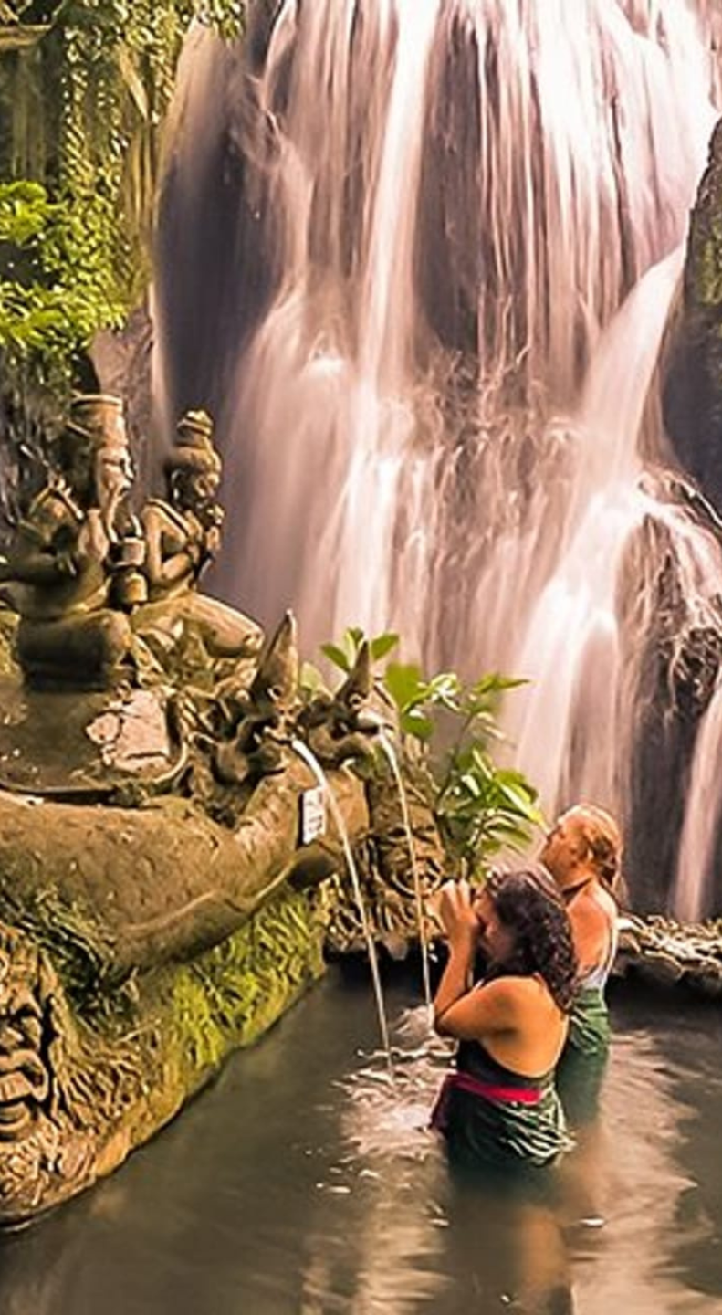People praying at a fountain with multiple small statues and a waterfall in the background.