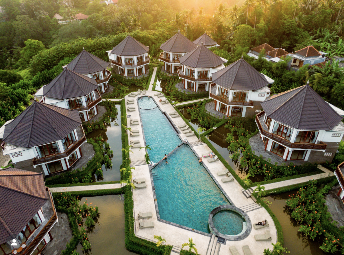 Aerial view of a tropical resort with multiple bungalow-style villas surrounding a large swimming pool and lush greenery at sunset.
