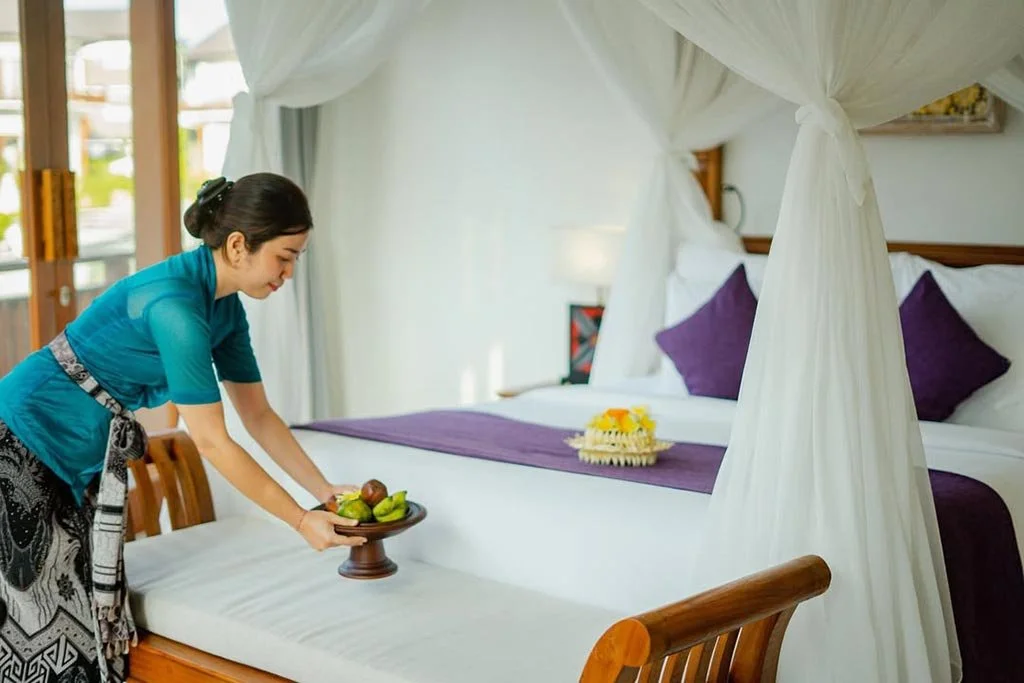 A woman in a teal top and patterned skirt arranges a tray of fresh fruit on a bed with a white canopy in a bright hotel room.