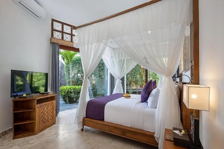 A bedroom with a wooden four-poster bed draped with white curtains, purple and white bedding, and a view of a lush garden through sliding glass doors. There's a wooden TV stand with a flat-screen TV, and a nightstand with a modern lamp.