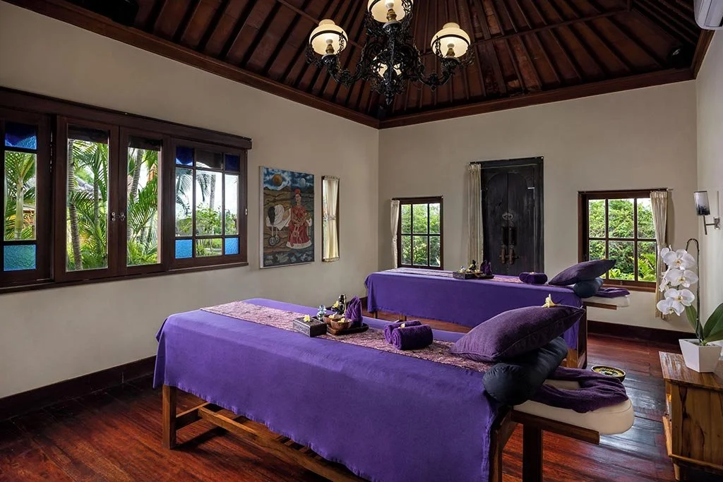 Two massage tables with purple sheets and pillows in a room with wooden floors, large windows, and a chandelier hanging from the wooden ceiling. There is a painting on the wall and a small table with a potted orchid plant.