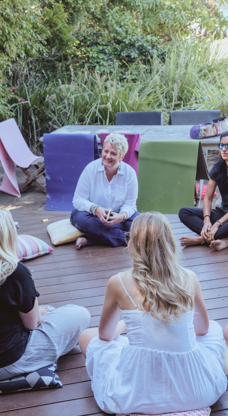 Group of people sitting outdoors on a wooden deck, engaged in conversation, with lush green plants in the background.