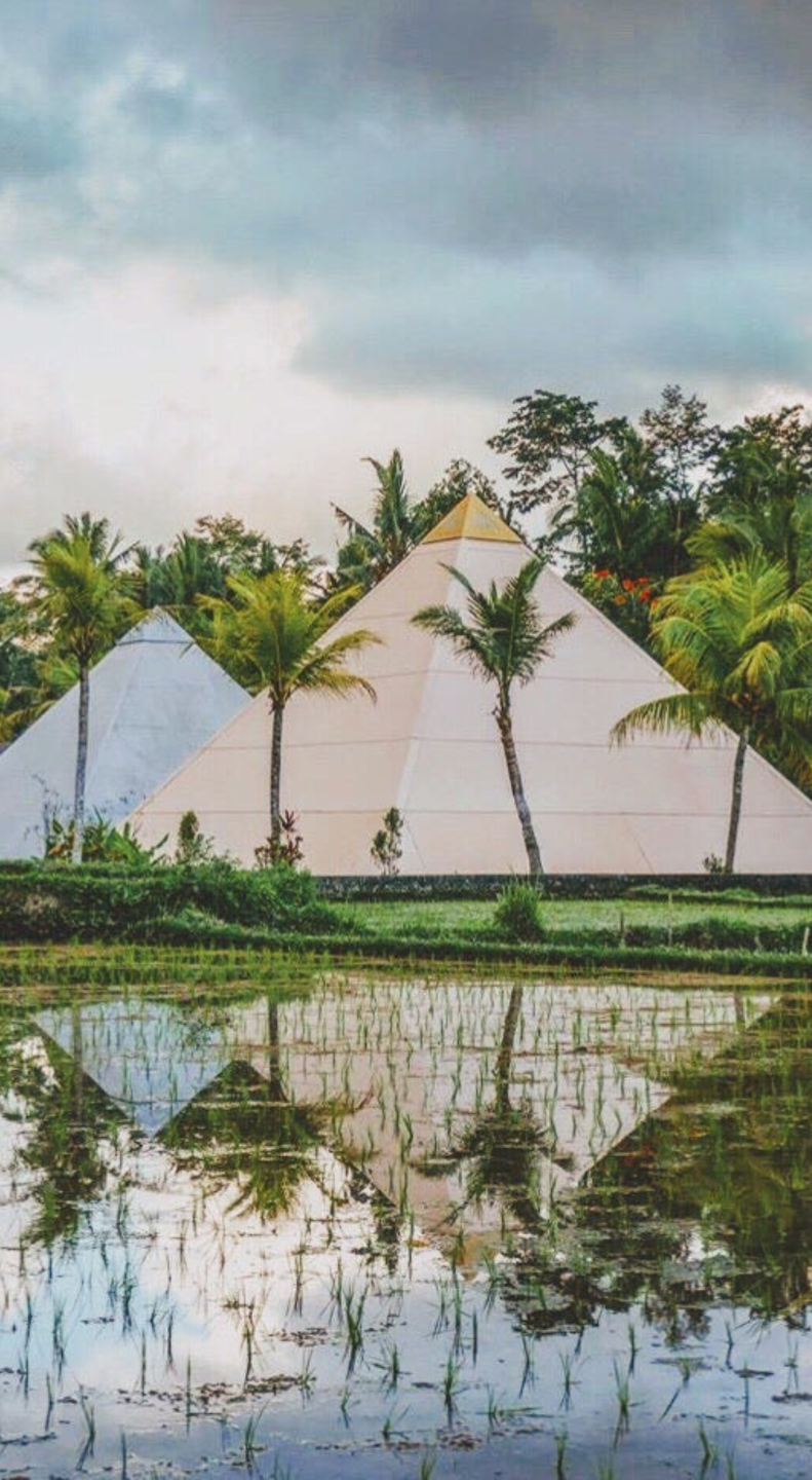 Large pyramid-shaped tent with a gold top, surrounded by palm trees, with a reflection in a pond in the foreground, and dark clouds in the sky.