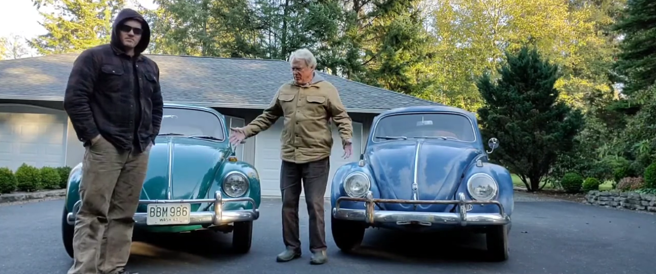 Two men standing on a driveway with two vintage Volkswagen Beetle cars parked behind them, surrounded by trees and a garage in the background.