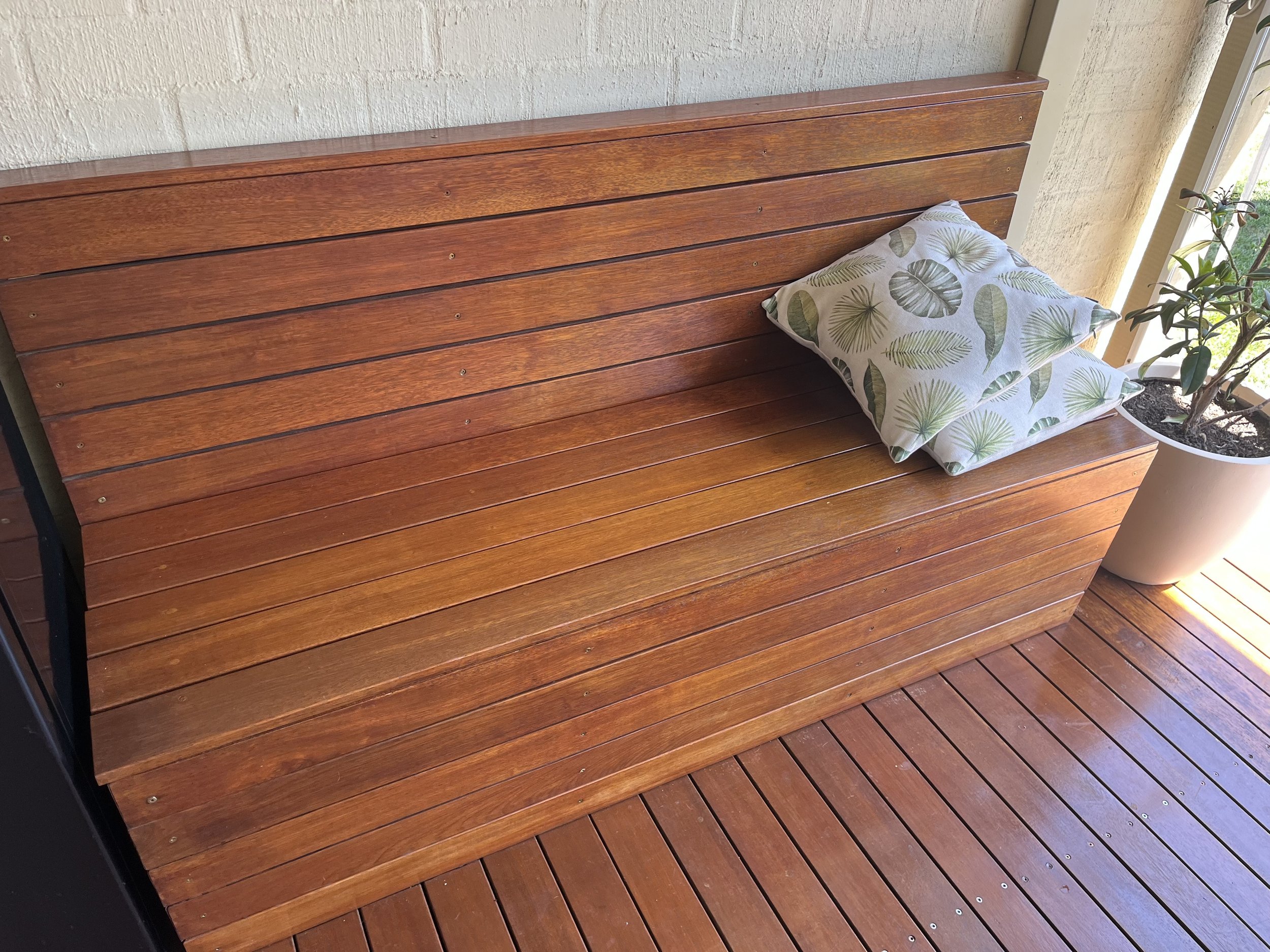 A wooden outdoor bench with horizontal slats and a patterned pillow, next to a potted plant, near a sliding glass door.