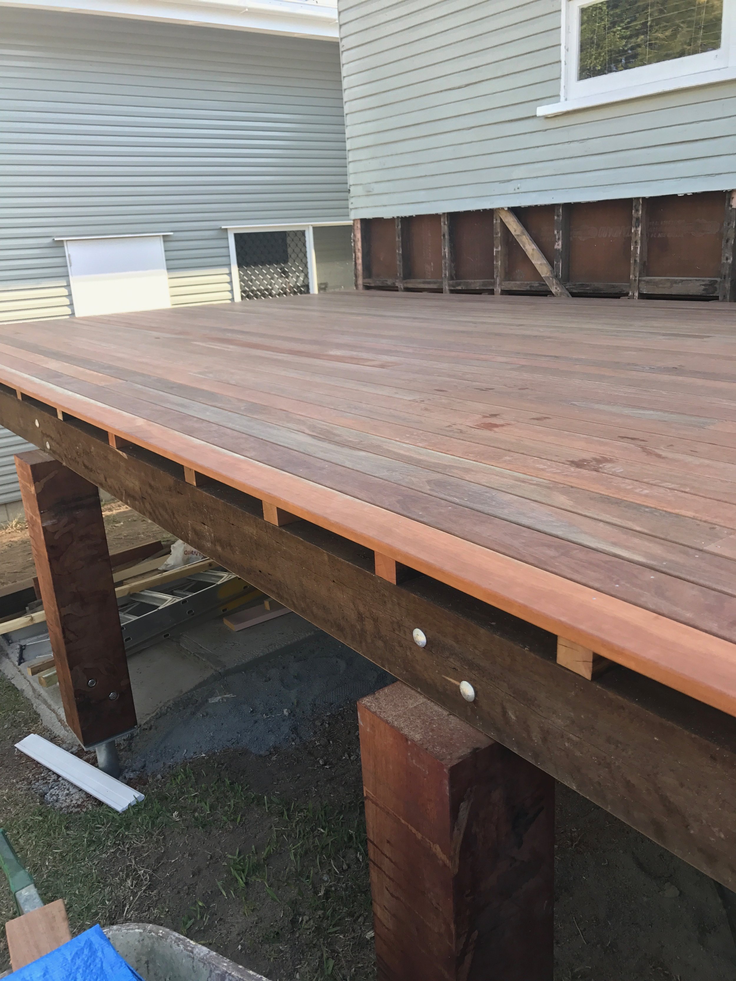 A wooden deck under construction next to a house with white siding. The deck is supported by large wooden posts, and the surface has wooden planks installed. Part of the house with a window is visible, and there are tools and materials around the construction site.