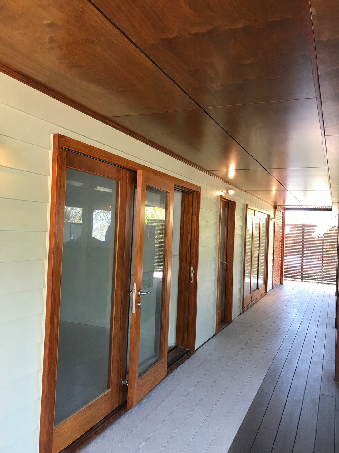 A covered outdoor corridor with glass doors and wooden frames leading to indoor rooms, with a wooden ceiling and a wooden privacy screen at the end.