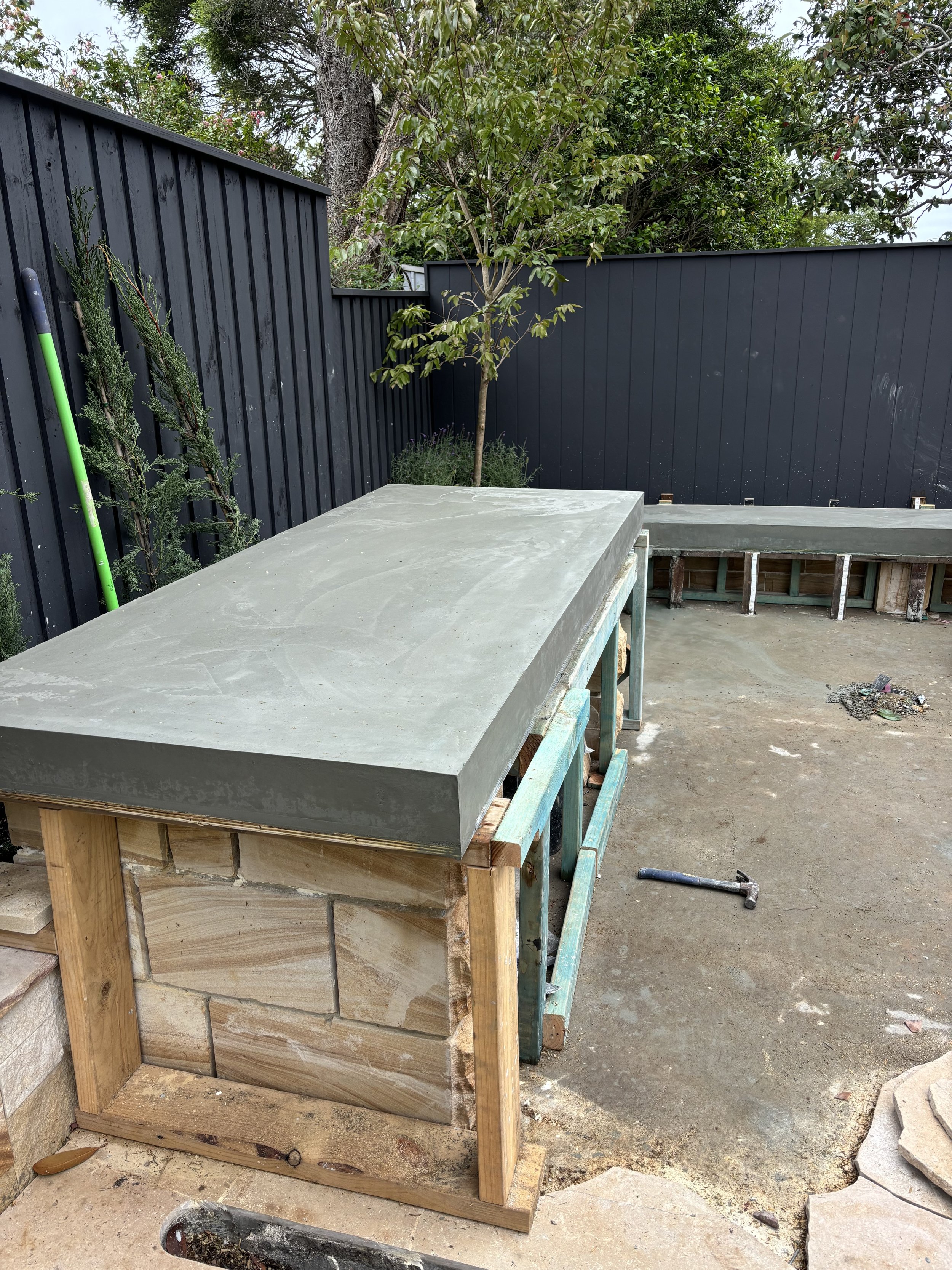 A construction site showing a concrete structure with wooden framing and a green tool leaning against a dark metal fence. There are trees and a patch of grass in the background.