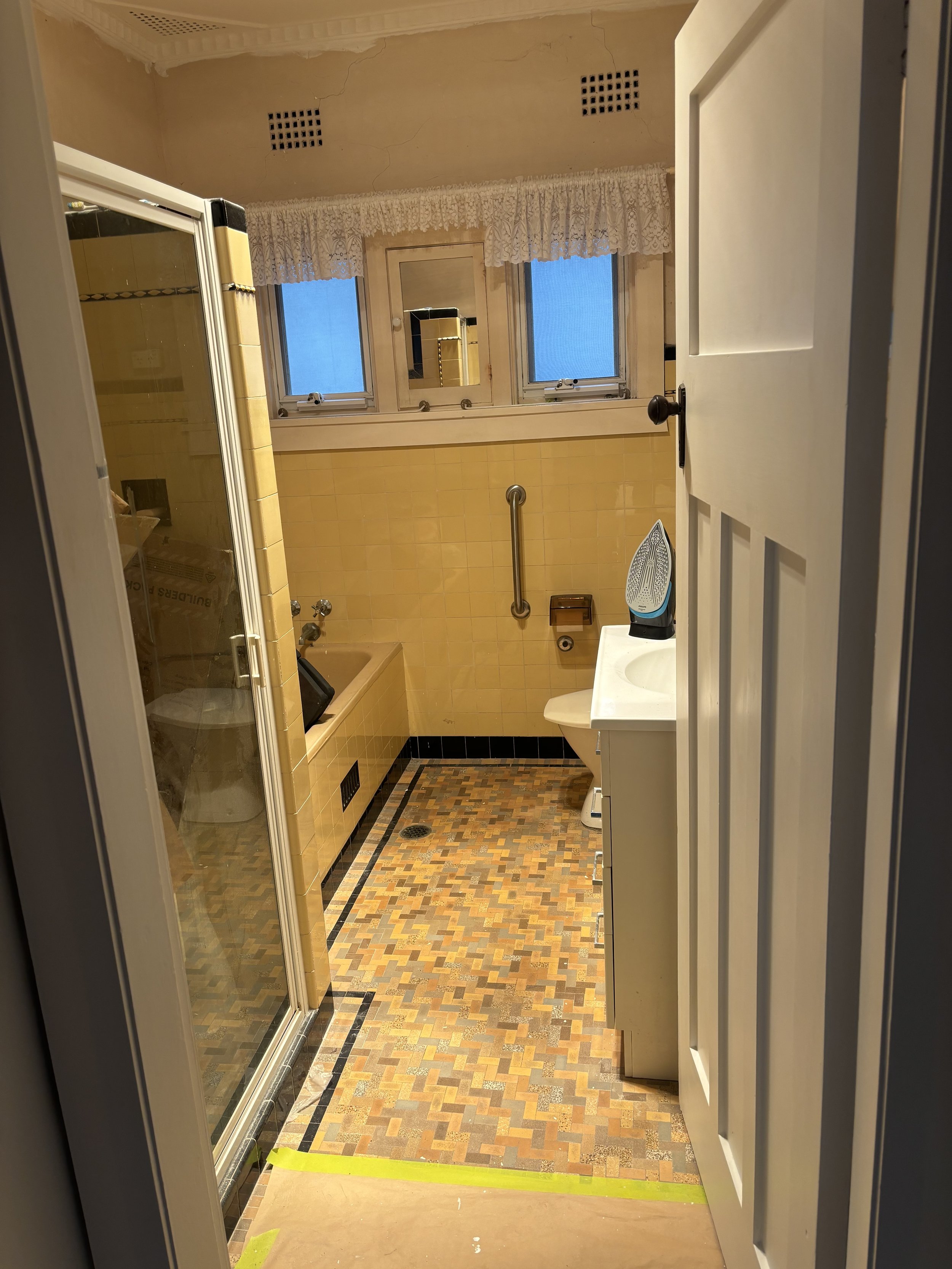 A view from a doorway into a bathroom with yellow tiles and a broad window with lace curtains, a mirror, a small white sink with items on it, and a patterned multicolored tile floor.