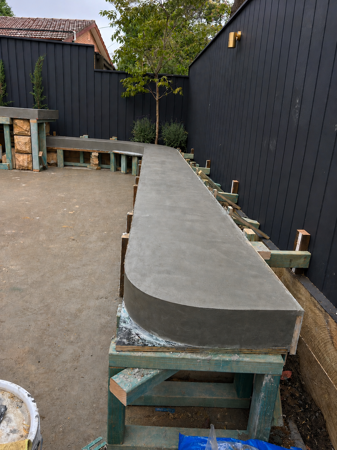 A backyard with a black fence, a potted tree, and plants. There is a curved concrete pathway under construction, supported by wooden frames. Construction materials and tools are visible nearby.