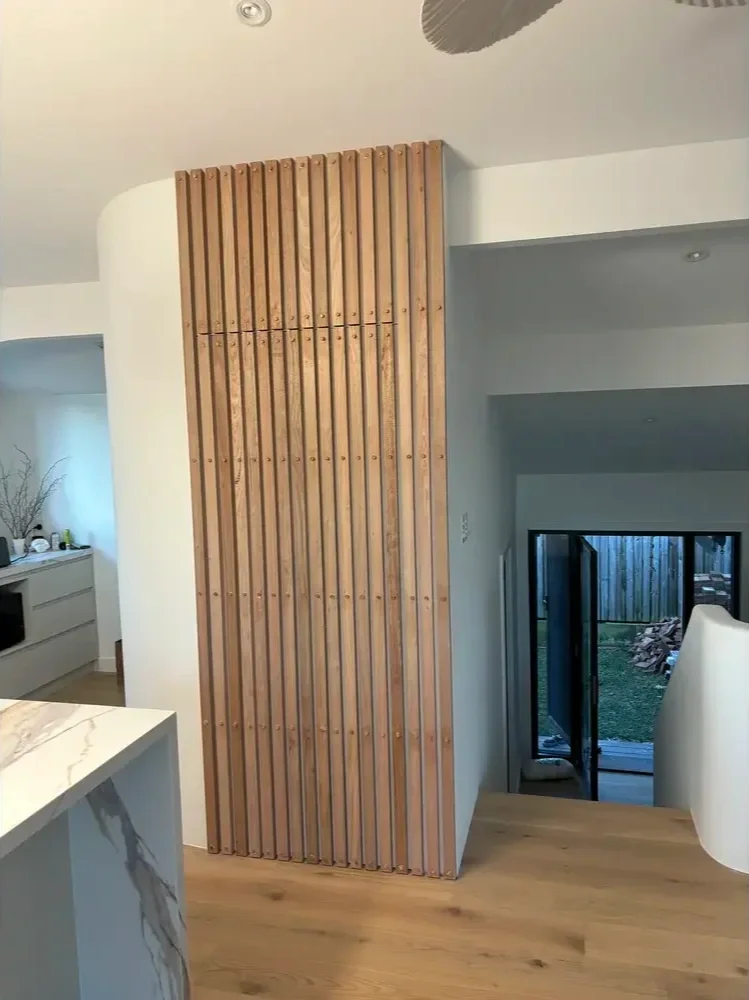 Interior view of a modern home featuring a vertical wooden slat wall, white ceiling, sliding glass door leading to backyard, and part of a kitchen with a marble countertop.