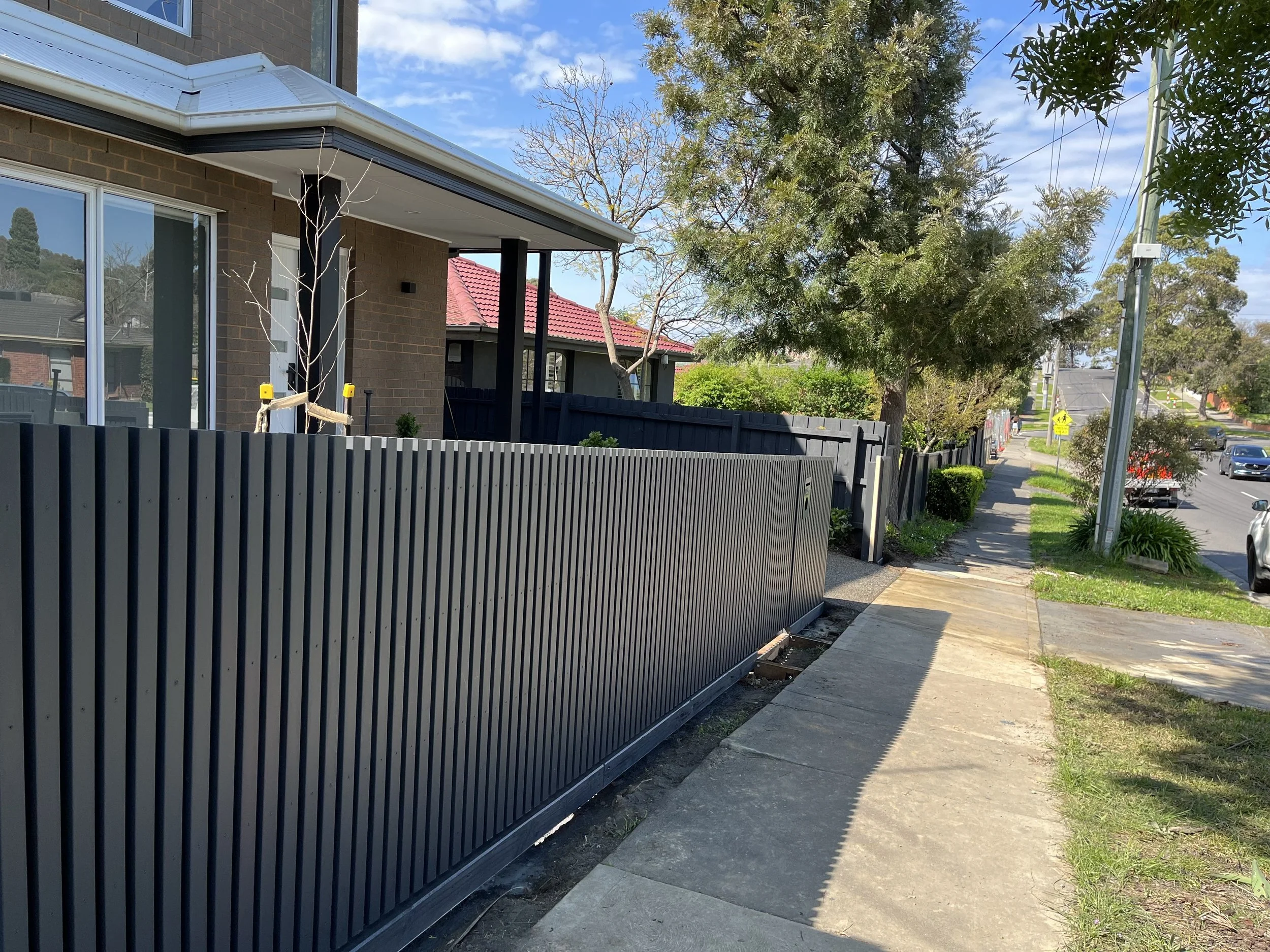 A sidewalk next to a black metal fence runs alongside a modern brick house with large windows. Trees and shrubs are visible, with one young, leafless tree tied to a support. A yellow pedestrian crossing sign is in the distance, with cars on the street and power lines overhead.
