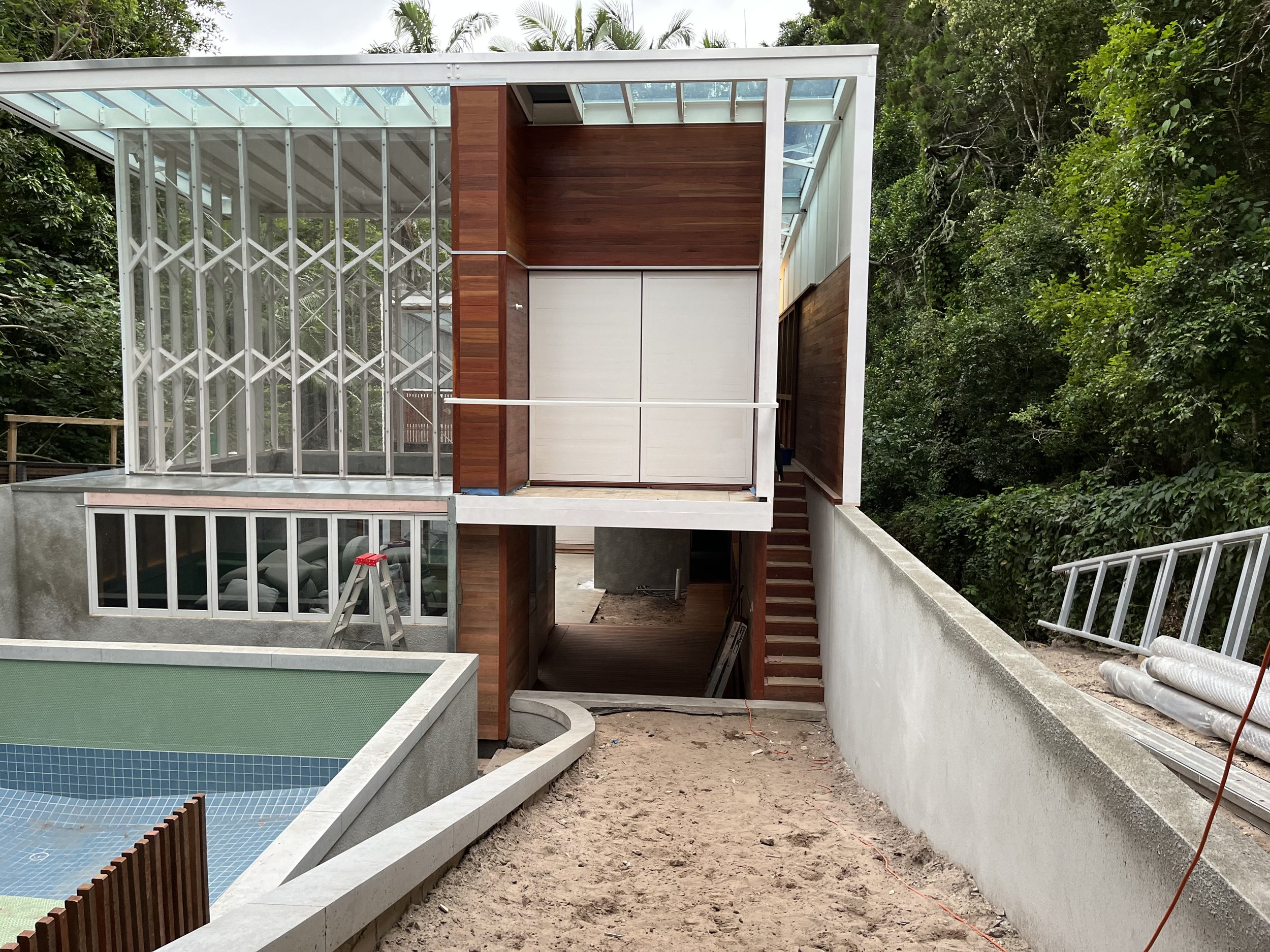A house under construction with a modern architectural design, featuring glass walls, wooden accents, and an open patio area, surrounded by greenery.