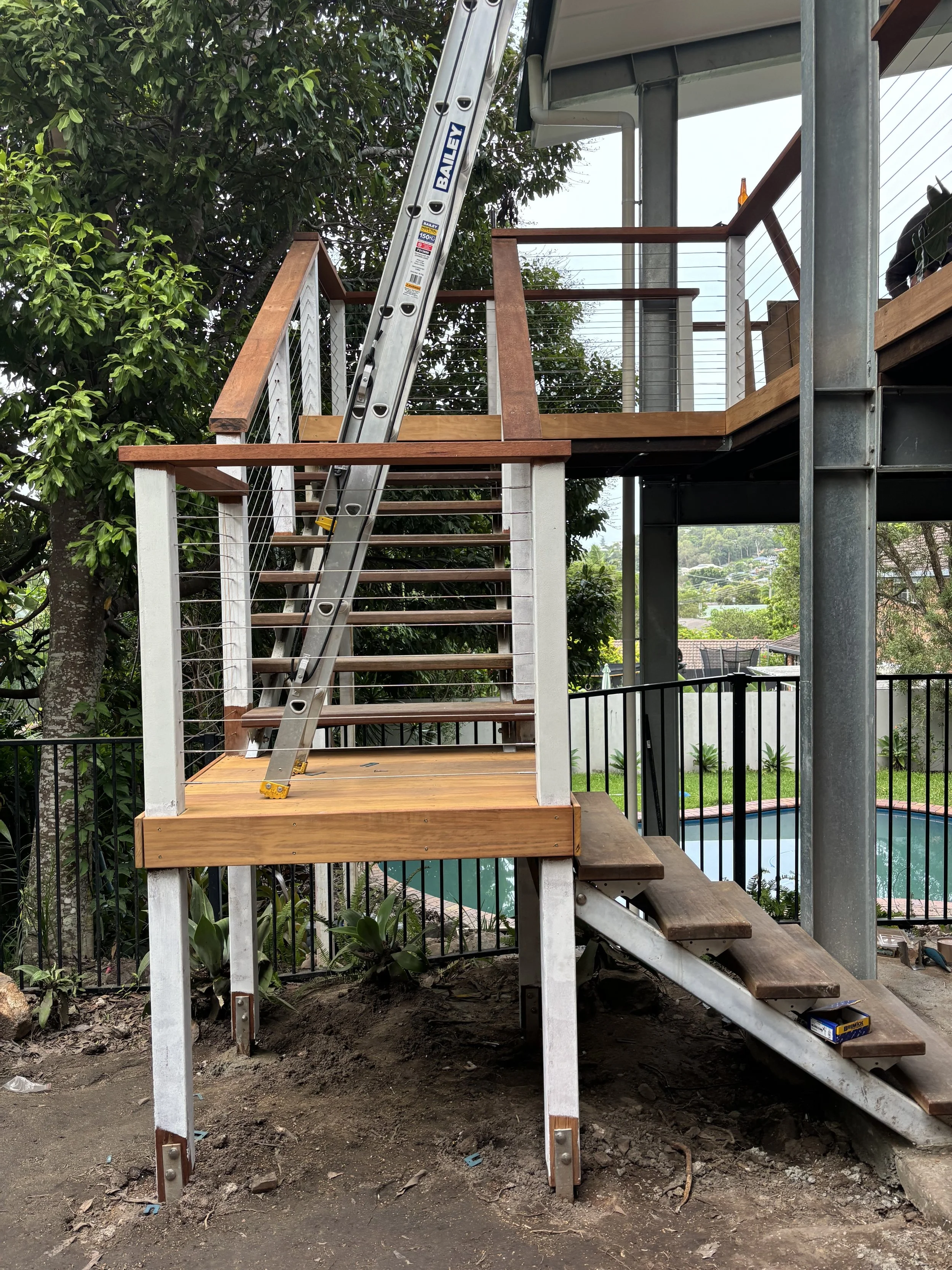 Construction site with wooden stairs, metal railing, a ladder leaning against the stairs, trees, and a swimming pool in the background.
