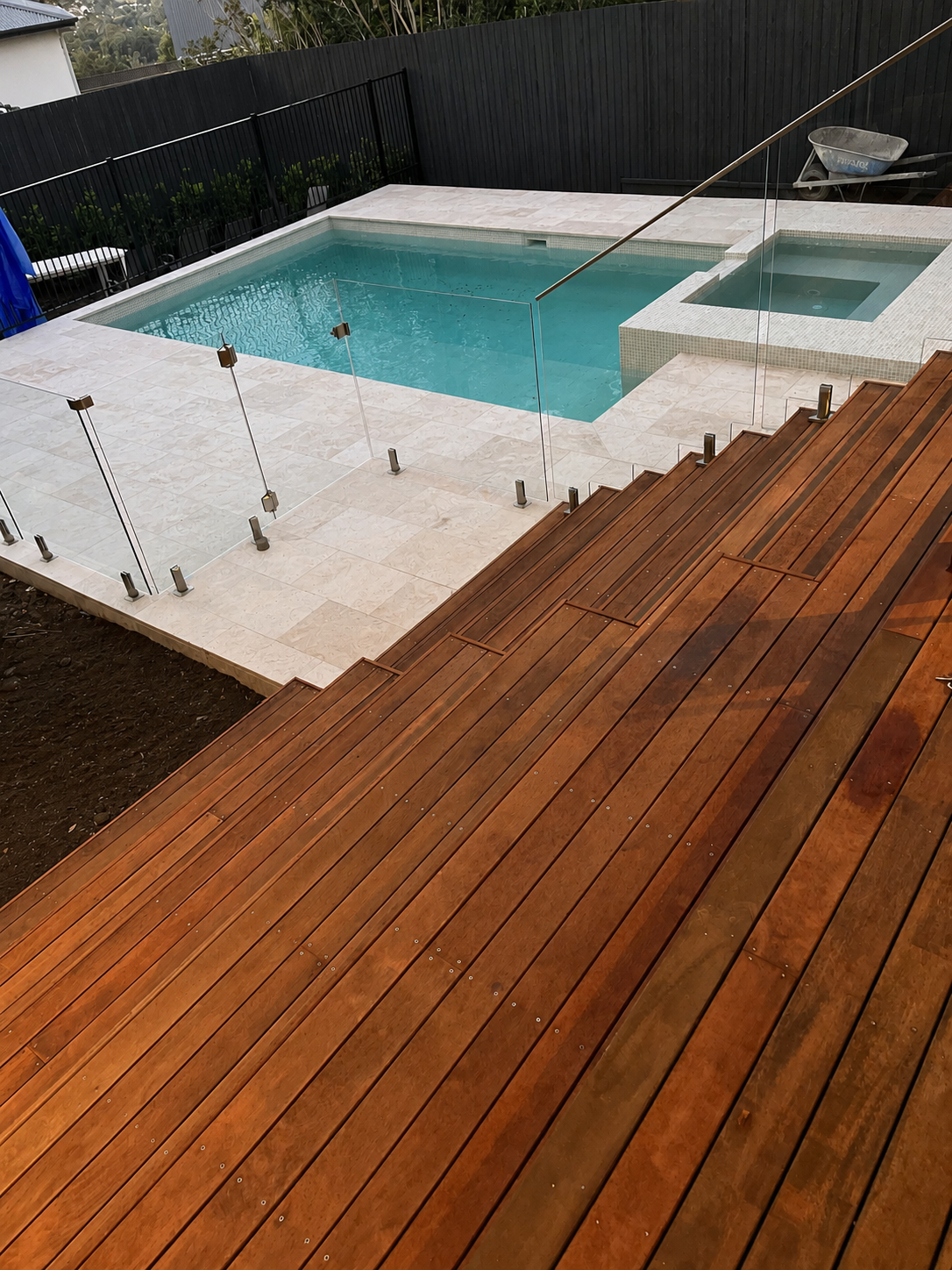A backyard with a swimming pool, a hot tub, a wooden deck, and a glass safety fence around the pool.