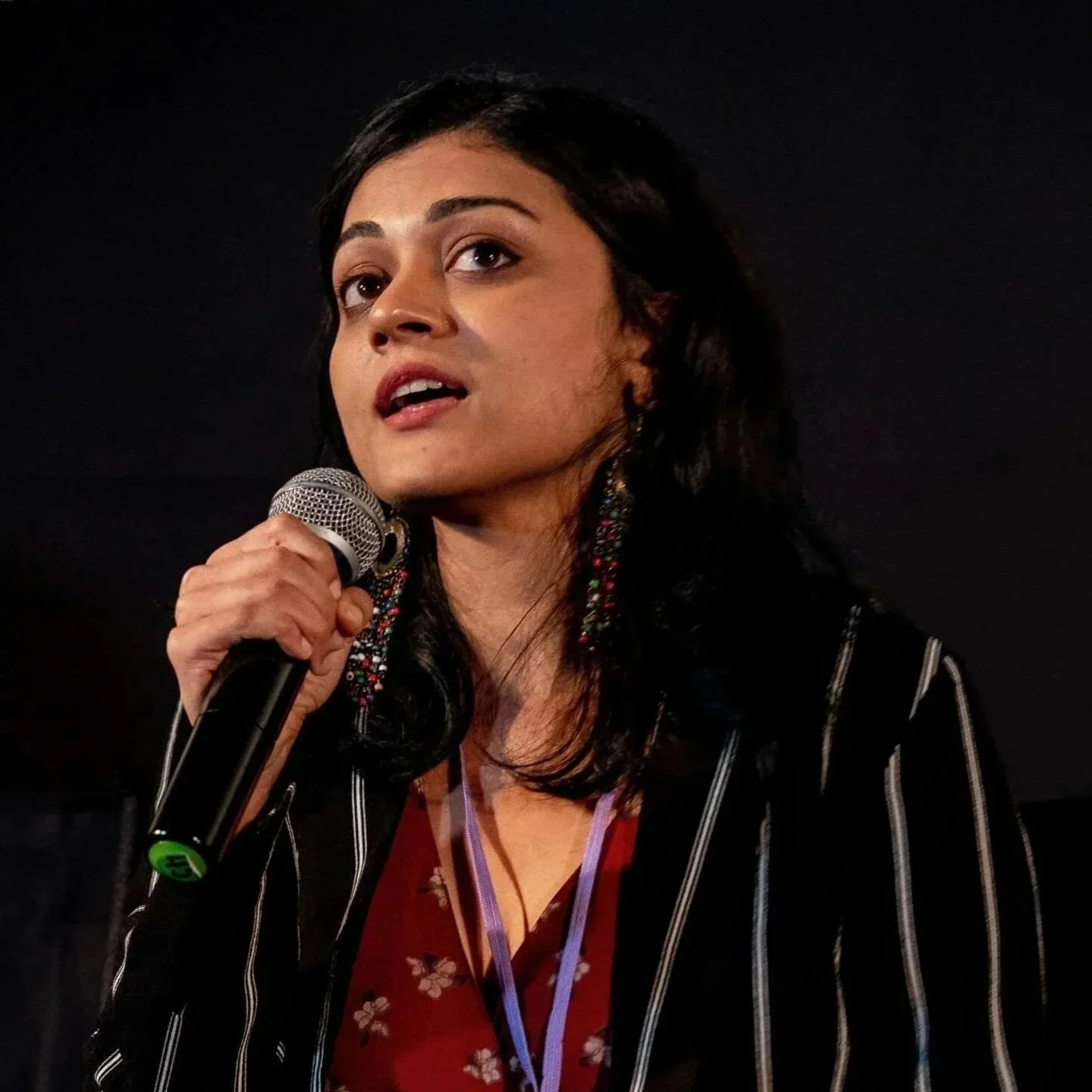 A woman with dark hair and earrings holding a microphone, speaking on stage with a dark background.