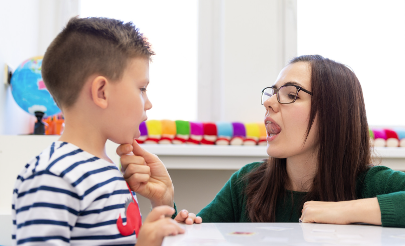 A female speech pathologist with long brown hair is modelling how to move her tongue to touch the roof of her mouth, and supporting a young boy to do the same. This is part of an oral motor exam