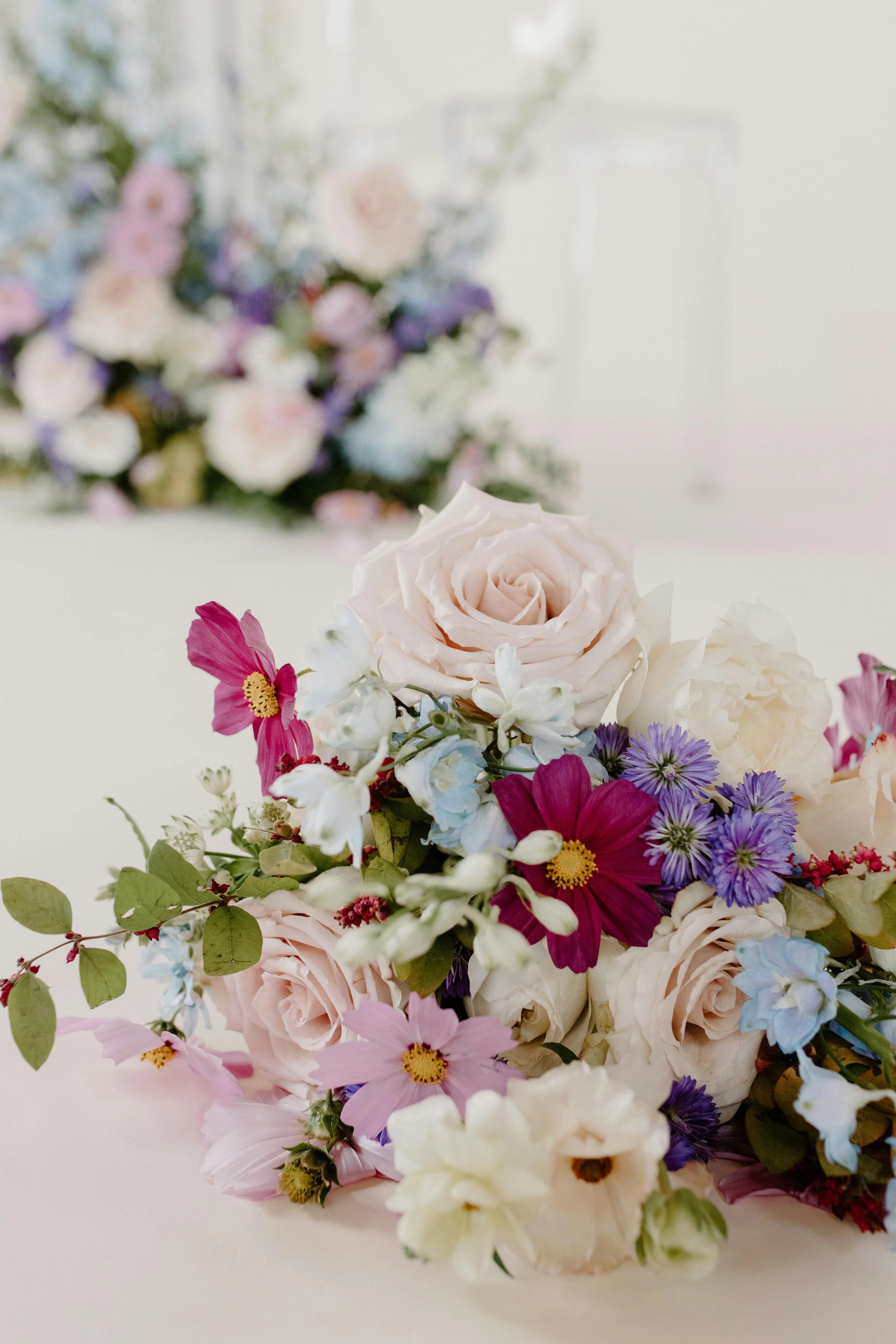 A close-up of a colorful floral arrangement with white roses, pink and purple flowers, and green leaves, set against a blurred background.