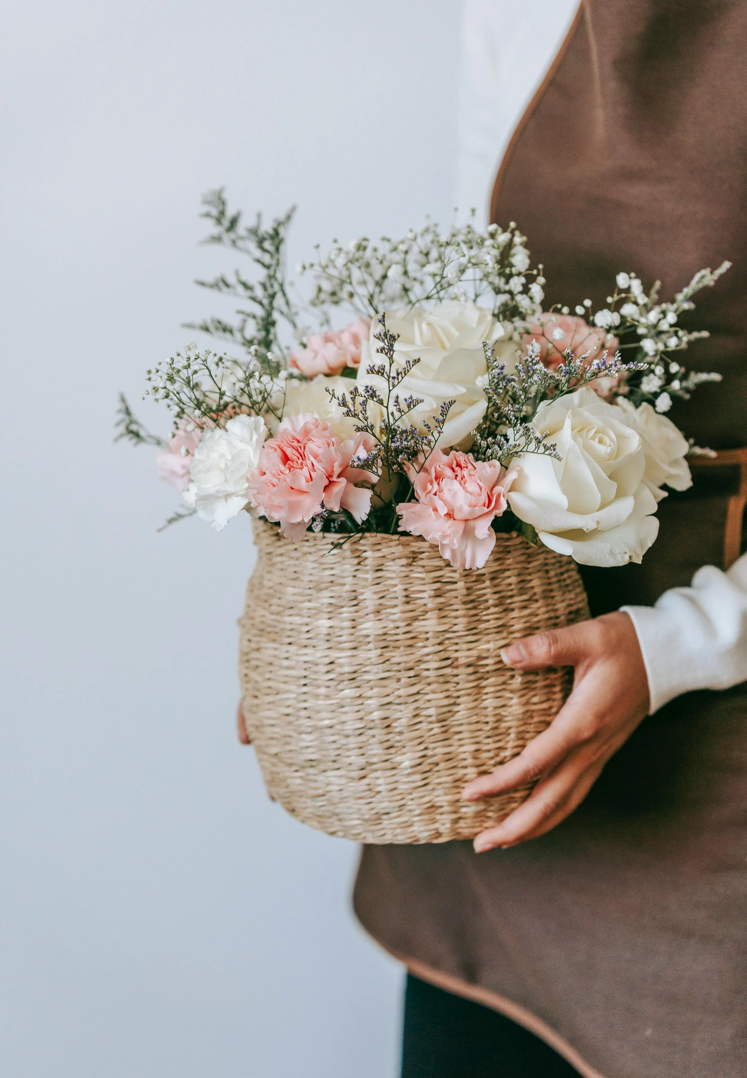 Person holding a woven basket filled with white roses, pink carnations, and small white and purple flowers.