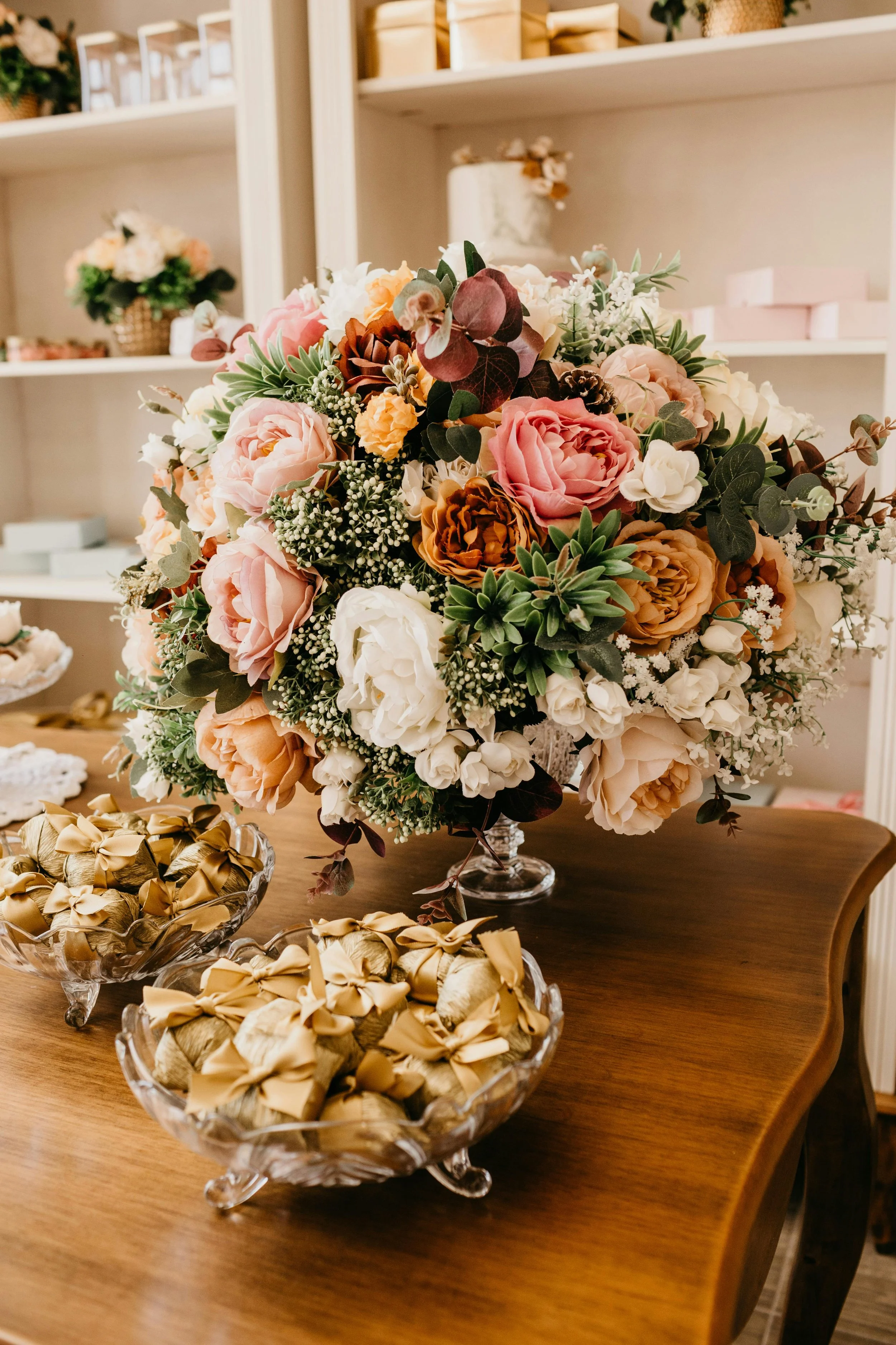 A large bouquet of mixed flowers including roses and succulents on a wooden table, with bowls of wrapped chocolates nearby.