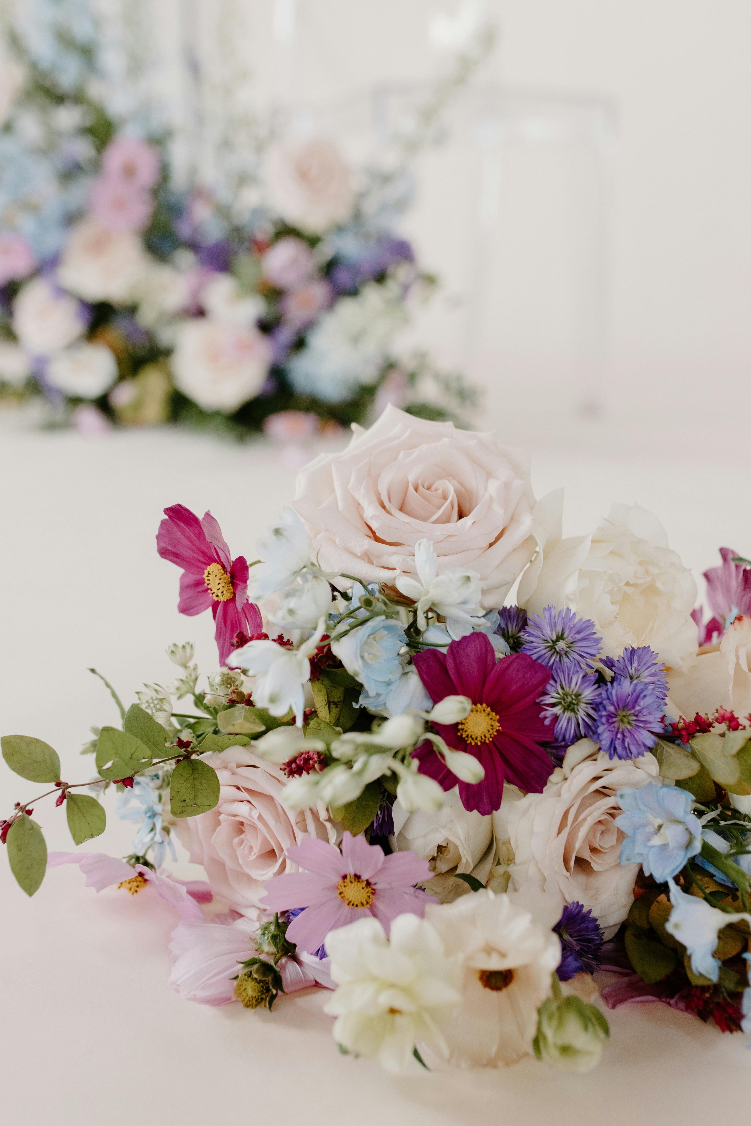 Close-up of a beautiful bouquet of flowers with roses, daisies, and other blooms in soft pastel and vibrant colors on a white surface, with a larger floral arrangement blurred in the background.