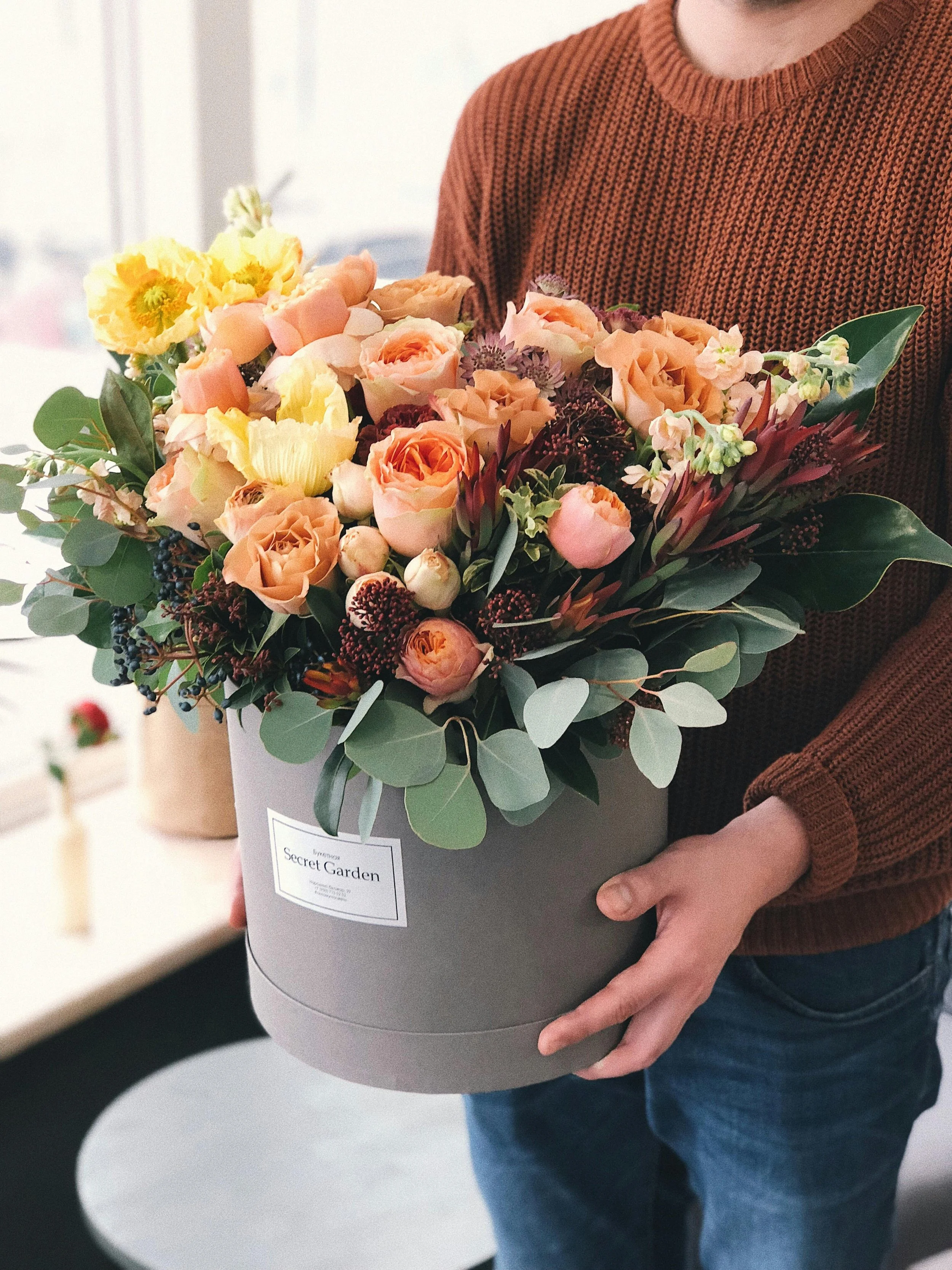 Person holding a large round gray box filled with a colorful bouquet of peach, pink, and yellow flowers with green foliage, labeled 'Secret Garden'.