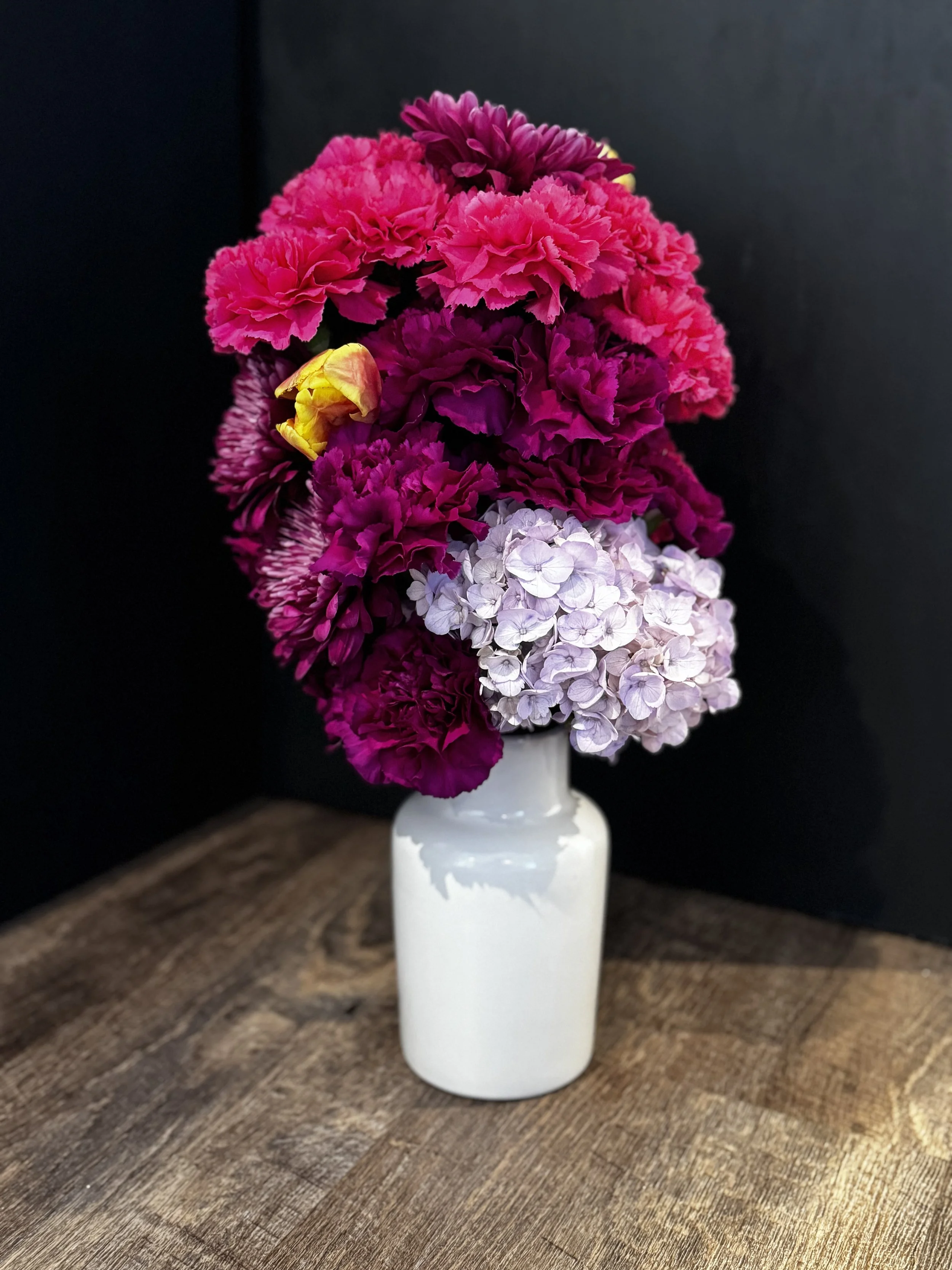 Concierge Desk Statement Arrangement — fuchsia carnations, plum mums, and a soft lavender hydrangea accent styled in a glossy white vessel for a bold, welcoming lobby moment.