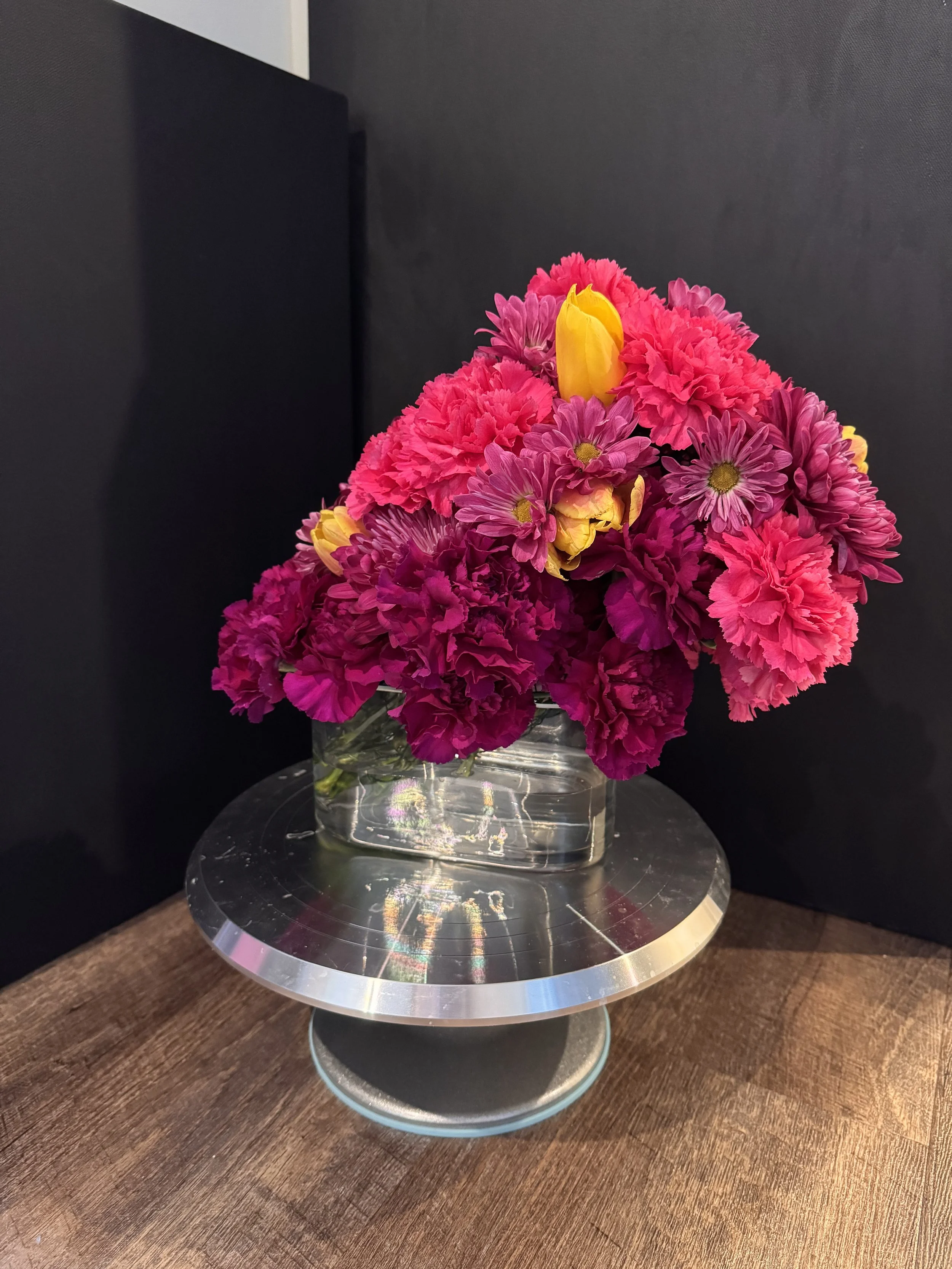 Host Stand / Bar Accent (Front View) — a compact jewel-tone arrangement in a modern clear cube with magenta and plum blooms finished with a golden tulip highlight.