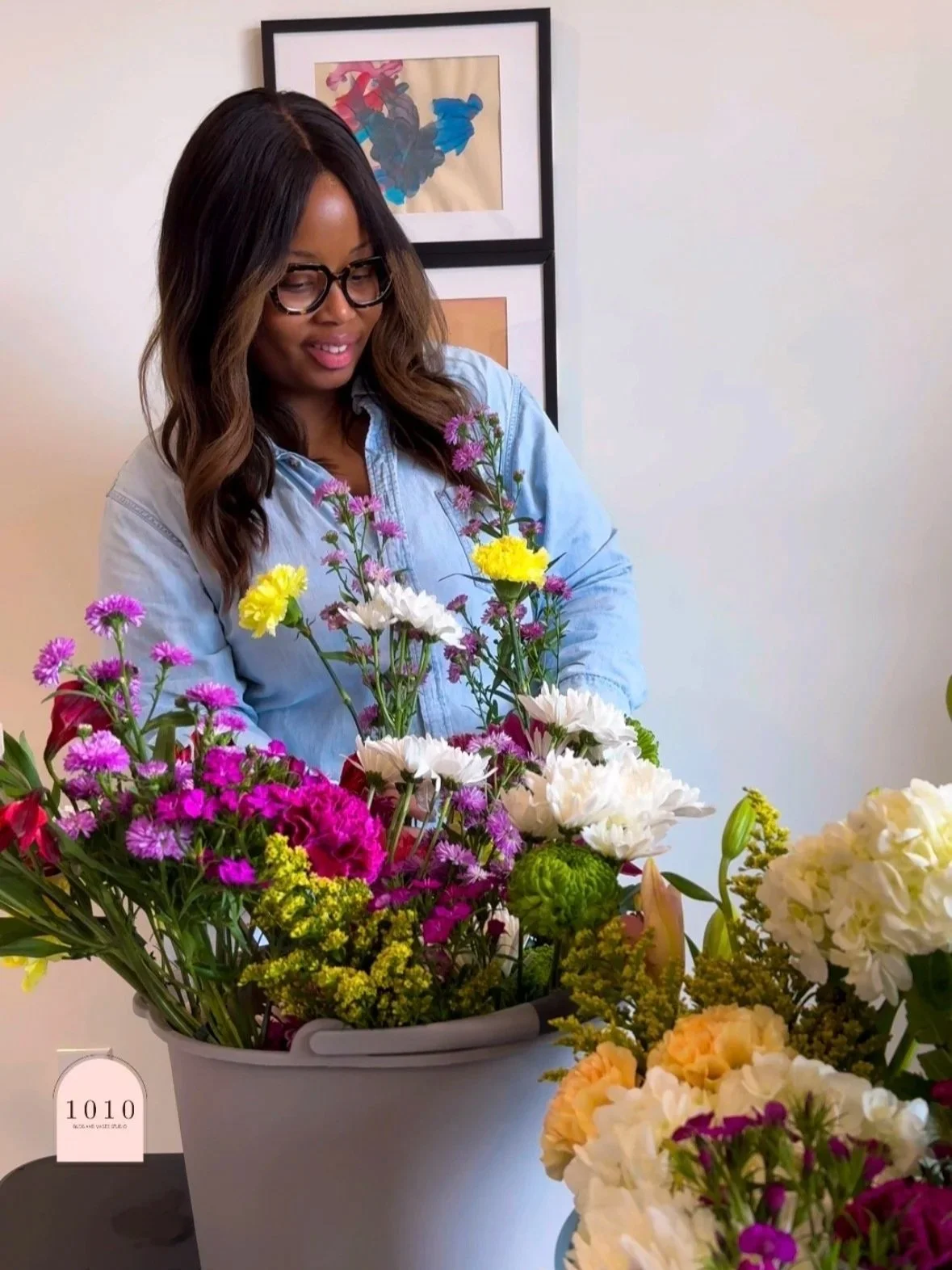 A woman with long wavy hair, wearing glasses and a light blue denim shirt, looking at a large basket of colorful flowers on a table. There are framed abstract art pieces on the wall behind her.