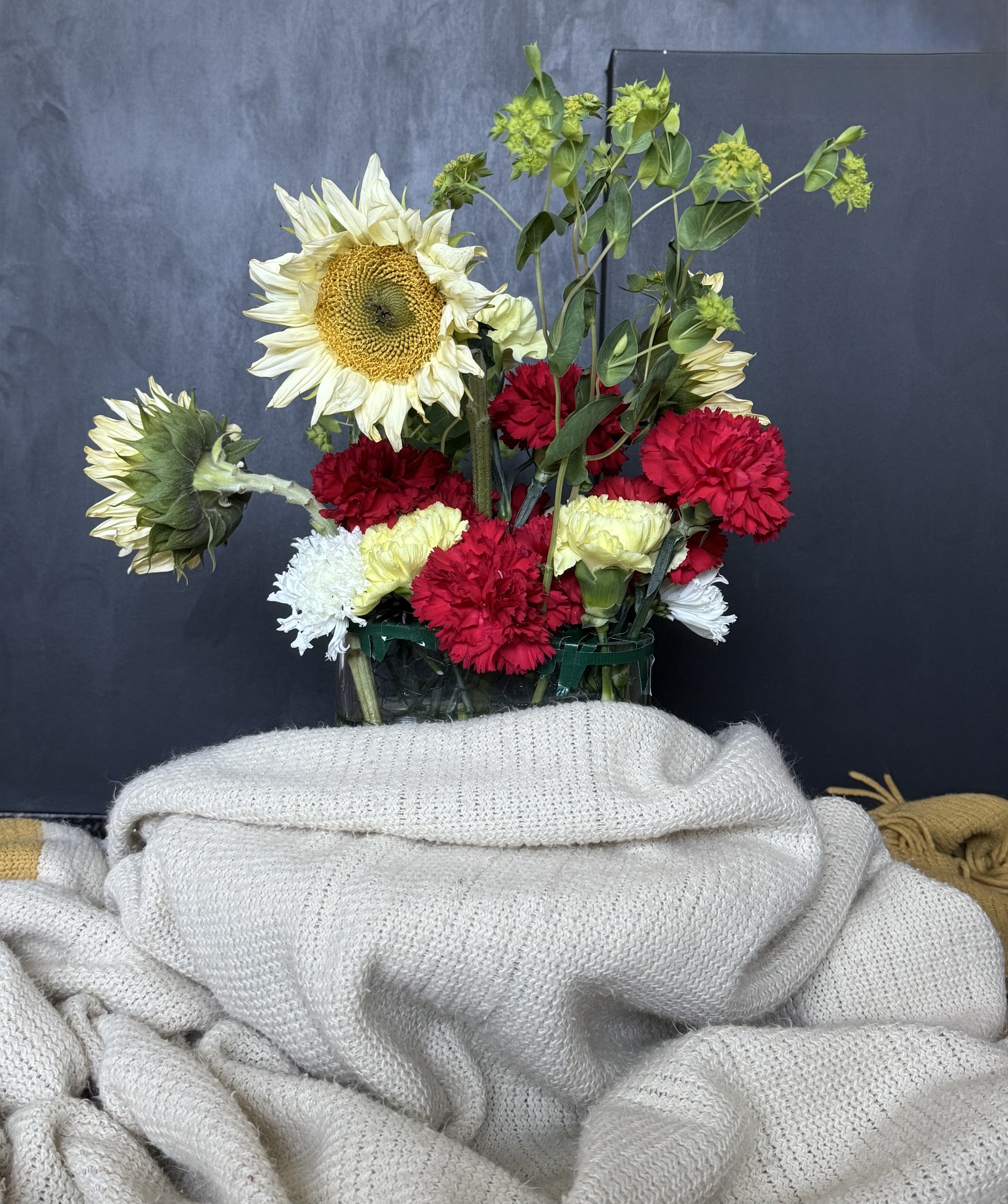  Lobby / Lounge Welcome Moment — sunflower-forward arrangement with crisp red carnations, soft buttercream accents, and airy greenery styled in a low clear vessel for an elevated, guest-facing feature.